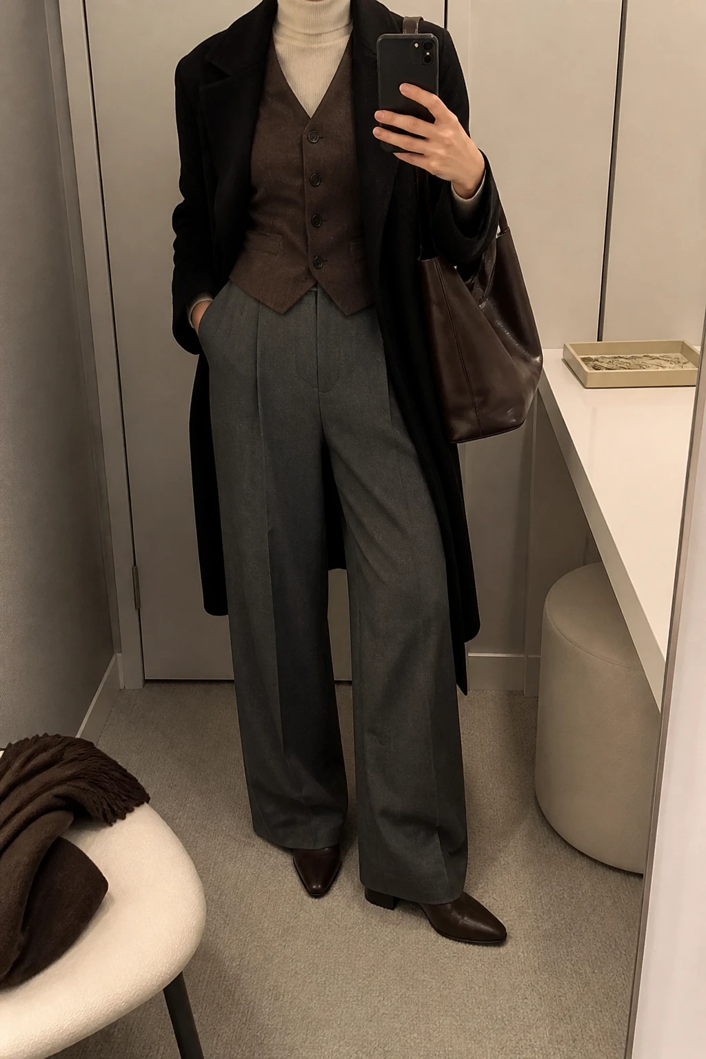 Image Prompt: Mirror selfie of a woman wearing a chocolate brown tailored wool waistcoat buttoned over a cream fitted fine-knit turtleneck sweater, charcoal grey high-waisted wide-leg tailored trousers, dark brown leather pointed-toe ankle boots with low block heels, a long black wool coat worn open, and a structured chocolate brown leather shoulder tote, holding a phone in front of a mirror, cropped at the NECK so the face does not show, with a relaxed casual selfie pose, in a minimal dressing room, soft indoor lighting, a folded blanket on a chair and a jewelry tray on the vanity, realistic chocolate wool waistcoat texture, natural grey tailored trouser folds and believable winter layering, latest iPhone photo quality, no additional clothing or accessories.