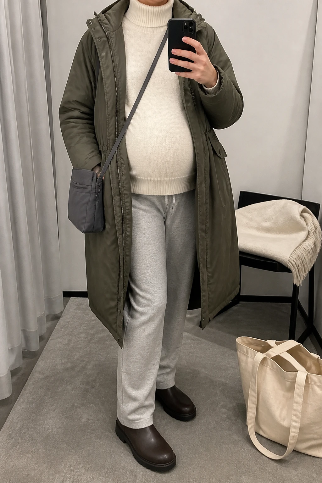 Image Prompt: Mirror selfie of a visibly pregnant woman wearing an olive green long insulated maternity parka with an adjustable expandable front panel, left open over a cream relaxed-fit thermal turtleneck sweater with room for her rounded belly, heather grey over-the-bump straight-leg soft knit maternity joggers, dark brown waterproof flat winter boots with rubber lug soles, and a small charcoal grey nylon crossbody bag, holding a phone in front of a mirror, cropped at the NECK so the face does not show, with a relaxed casual selfie pose and natural visible pregnancy silhouette, in a minimal dressing room, soft indoor lighting, a tote bag resting on the floor and a folded blanket on a chair, realistic olive parka fabric, natural grey knit jogger folds, believable maternity fit, latest iPhone photo quality, no additional clothing or accessories.