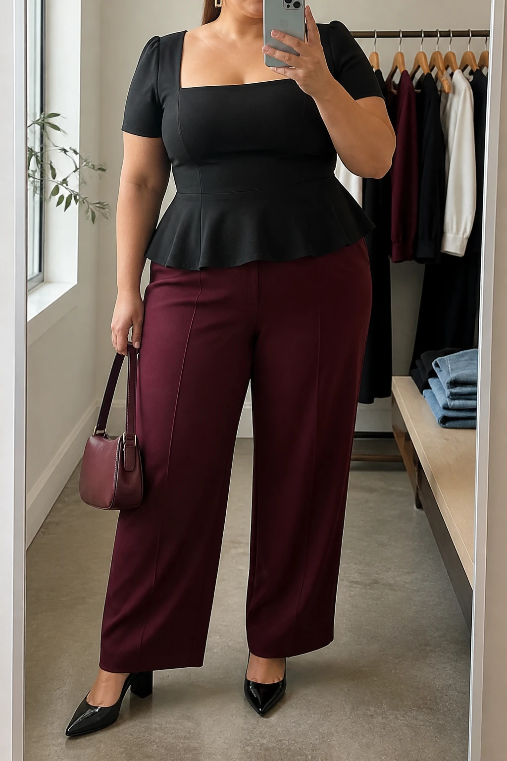Mirror selfie of a plus size woman wearing a structured black square-neck peplum top with short sleeves, high-waisted burgundy wide-leg trousers in fluid crepe, black pointed block heels, a small burgundy leather handbag, and gold rectangular earrings, clearly showing the exact clothing items, black peplum shape, burgundy trousers, colors, fabrics, footwear, and accessories listed, holding a phone in front of a mirror, cropped at the neck so her face does not show, relaxed confident selfie pose, in a minimal dressing room with natural window light, a clothing rack with garments behind her and folded denim jeans on a bench, realistic graduation outfit, latest iPhone photo quality.