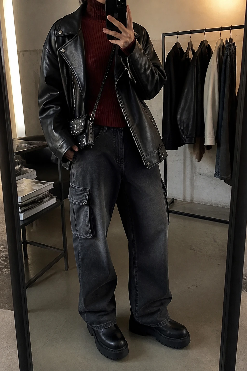 Image Prompt: Mirror selfie of a woman wearing an oversized black faux-leather biker jacket over a burgundy fitted ribbed turtleneck sweater, charcoal grey baggy denim cargo jeans with side pockets, black leather chunky platform combat boots with thick flat soles, and a small black studded leather crossbody bag, holding a phone in front of a mirror, cropped at the NECK so the face does not show, with a relaxed casual selfie pose, in a fashion studio mirror corner, warm moody indoor lighting, a clothing rack with garments behind and a stack of fashion magazines on a side table, realistic black leather texture, natural charcoal denim creases and edgy casual styling, latest iPhone photo quality, no additional clothing or accessories.