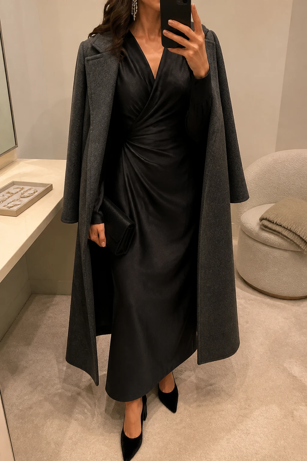 Image Prompt: Mirror selfie of a woman wearing a black long-sleeve satin maxi dress with a softly draped waist and modest V-neckline, black velvet pointed-toe block heels, long silver crystal drop earrings, a small black satin evening clutch, and a charcoal grey wool wrap coat worn open, holding a phone in front of a mirror, cropped at the NECK so the face does not show, with a relaxed casual selfie pose, in a minimal dressing room, warm soft indoor lighting, a jewelry tray on the vanity and a folded blanket on a chair, realistic flowing satin folds, soft reflections without artificial gloss, latest iPhone photo quality, no additional clothing or accessories.
