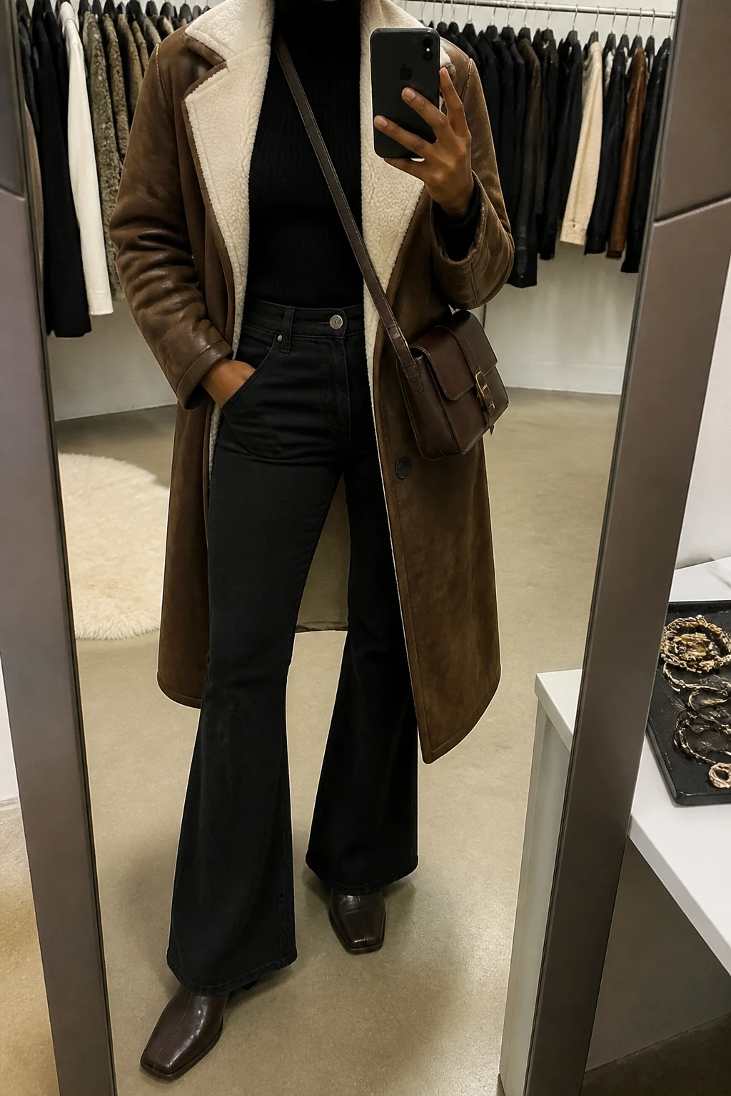 Image Prompt: Mirror selfie of a black woman wearing a long chocolate brown faux-suede shearling-lined coat with a cream fleece collar over a black fitted ribbed turtleneck sweater, black high-waisted flared denim jeans, dark brown leather square-toe low block-heel ankle boots, and a small dark brown leather crossbody bag, holding a phone in front of a mirror, cropped at the NECK so the face does not show, with a relaxed casual selfie pose, in a fashion studio mirror corner, warm indoor lighting, a clothing rack with garments behind and a jewelry tray on the vanity, believable faux-suede and shearling textures, natural denim folds, latest iPhone photo quality, no additional clothing or accessories.