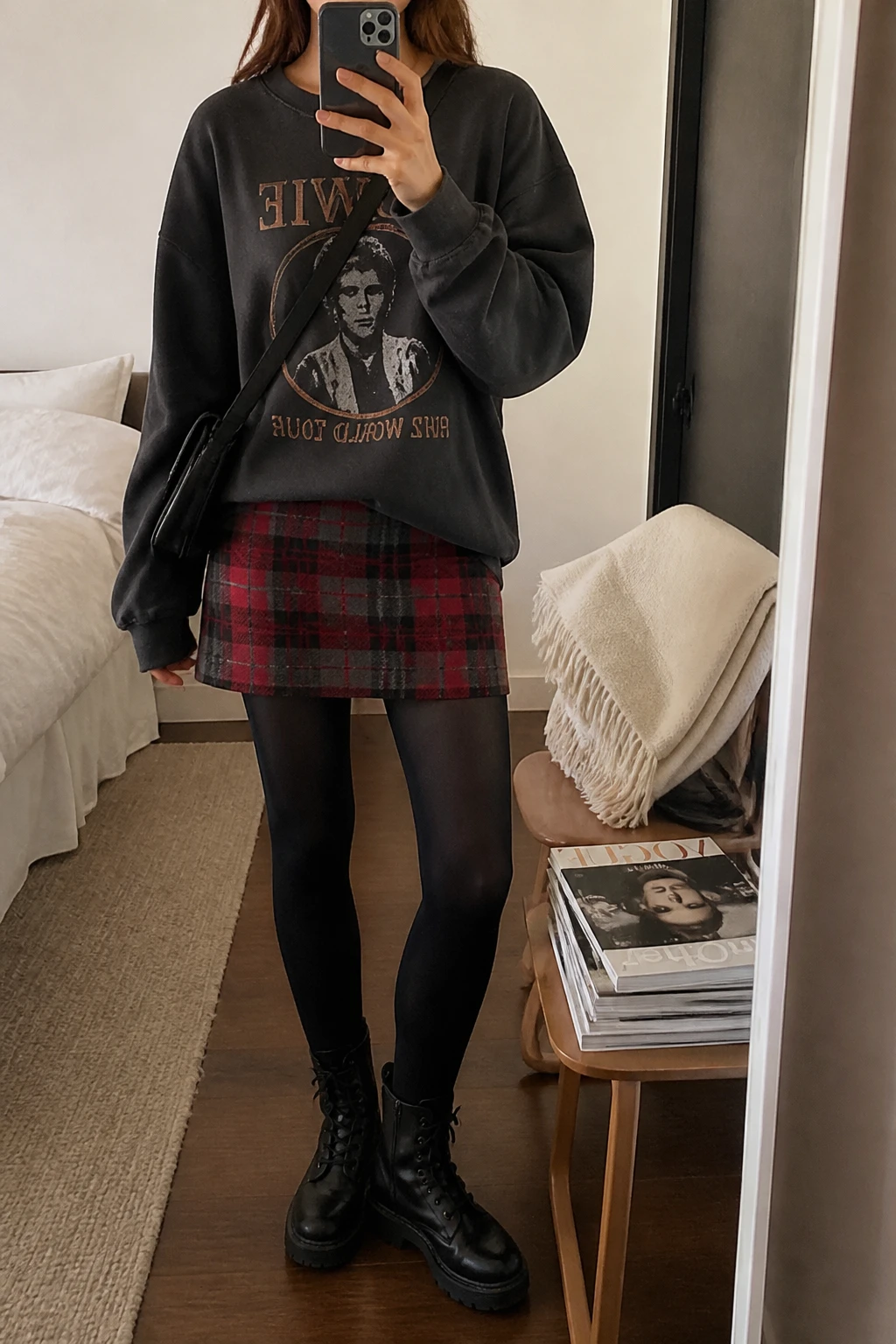 Image Prompt: Mirror selfie of a woman wearing an oversized washed black cotton graphic sweatshirt with a vintage music-inspired print, a red and charcoal plaid wool mini skirt, black opaque fleece-lined tights, black leather chunky lace-up combat boots, and a small black nylon crossbody bag, holding a phone in front of a mirror, cropped at the NECK so the face does not show, with a relaxed casual selfie pose, in a cozy bedroom mirror setup, soft indoor lighting, a folded blanket on a chair and a stack of fashion magazines on a side table, realistic cotton sweatshirt and plaid fabric, natural casual outfit folds, latest iPhone photo quality, no additional clothing or accessories.