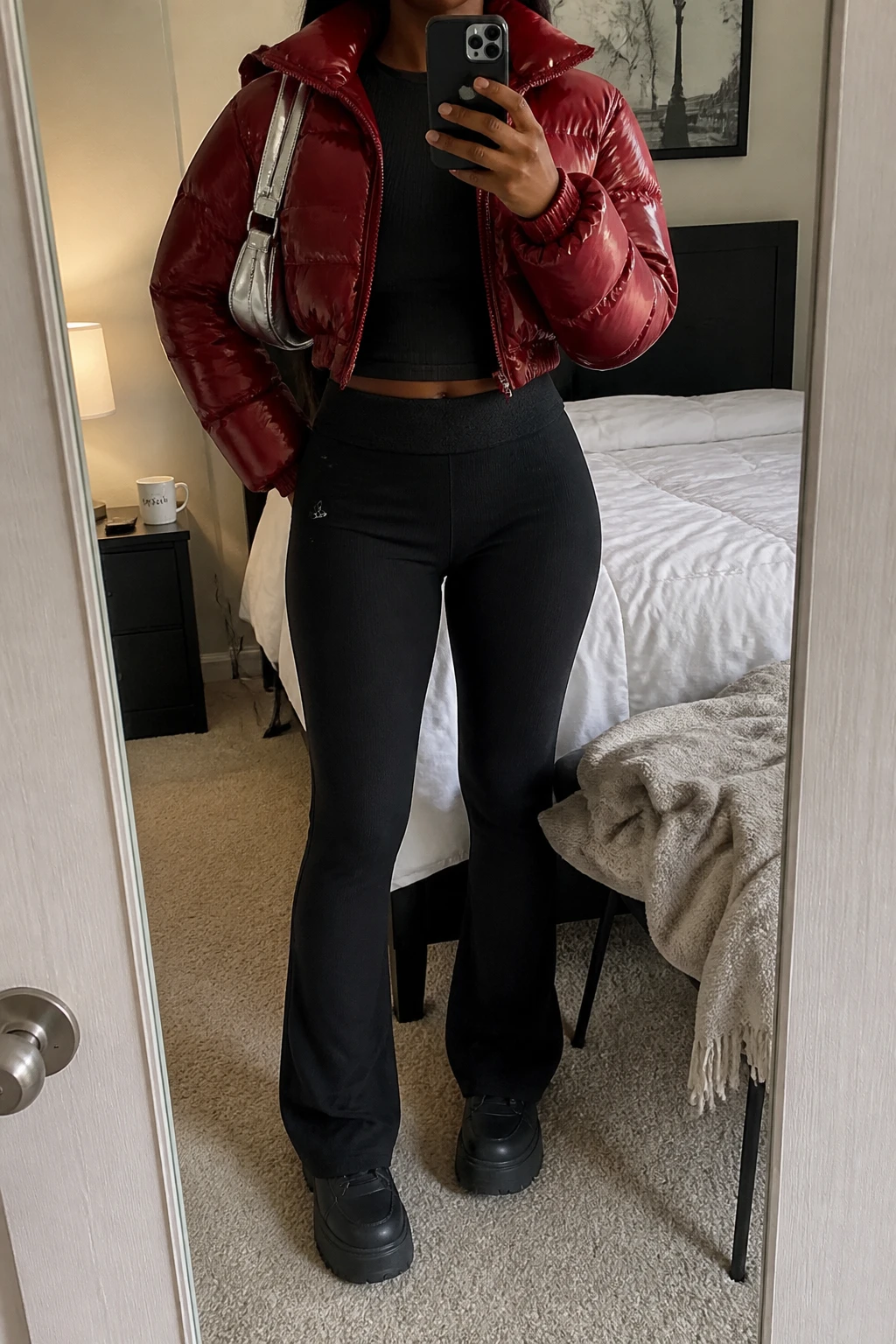 Image Prompt: Mirror selfie of a black woman wearing a cherry red cropped insulated puffer jacket over a black fitted ribbed long-sleeve mock-neck top, black high-waisted fleece-lined flared leggings, black platform leather sneakers with thick flat soles, and a small metallic silver nylon shoulder bag, holding a phone in front of a mirror, cropped at the NECK so the face does not show, with a relaxed casual selfie pose, in a cozy bedroom mirror setup, soft indoor lighting, a coffee mug on a bedside table and a folded blanket on a chair, natural cherry red puffer color, realistic winter fabric folds and early 2000s sporty styling, latest iPhone photo quality, no additional clothing or accessories.