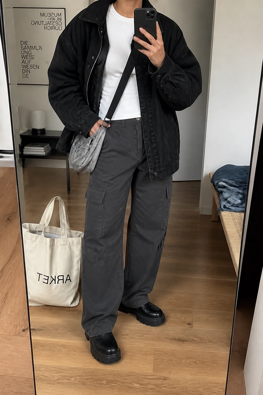 Image Prompt: Mirror selfie of a woman wearing an oversized black denim jacket with a warm quilted lining over a fitted white heavyweight cotton crewneck top, charcoal grey straight-leg cargo trousers with side pockets, black leather chunky platform combat boots with flat rubber soles, and a small silver-grey nylon crossbody bag, holding a phone in front of a mirror, cropped at the NECK so the face does not show, with a relaxed casual selfie pose, in a stylish city apartment, soft indoor lighting, a tote bag resting on the floor and folded denim jeans on a bench, believable cargo fabric texture, natural jacket folds, latest iPhone photo quality, no additional clothing or accessories.