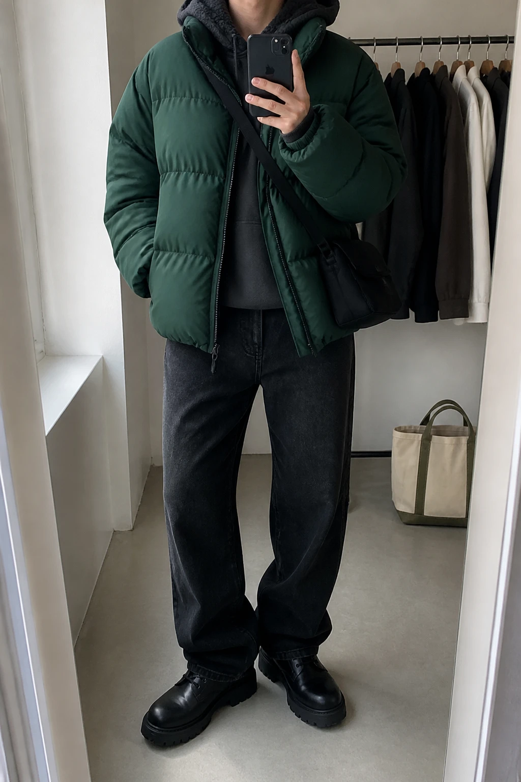 Image Prompt: Mirror selfie of a woman wearing a forest green oversized insulated quilted puffer jacket over a charcoal grey thick fleece-lined hoodie, washed black baggy straight-leg denim jeans, black waterproof leather chunky lace-up boots with flat rubber soles, and a small black nylon crossbody bag, holding a phone in front of a mirror, cropped at the NECK so the face does not show, with a relaxed casual selfie pose, in a minimal dressing room, soft natural window light, a tote bag resting on the floor and a clothing rack with garments behind, realistic forest green puffer volume, natural grey hoodie folds and relaxed black denim creases, latest iPhone photo quality, no additional clothing or accessories.