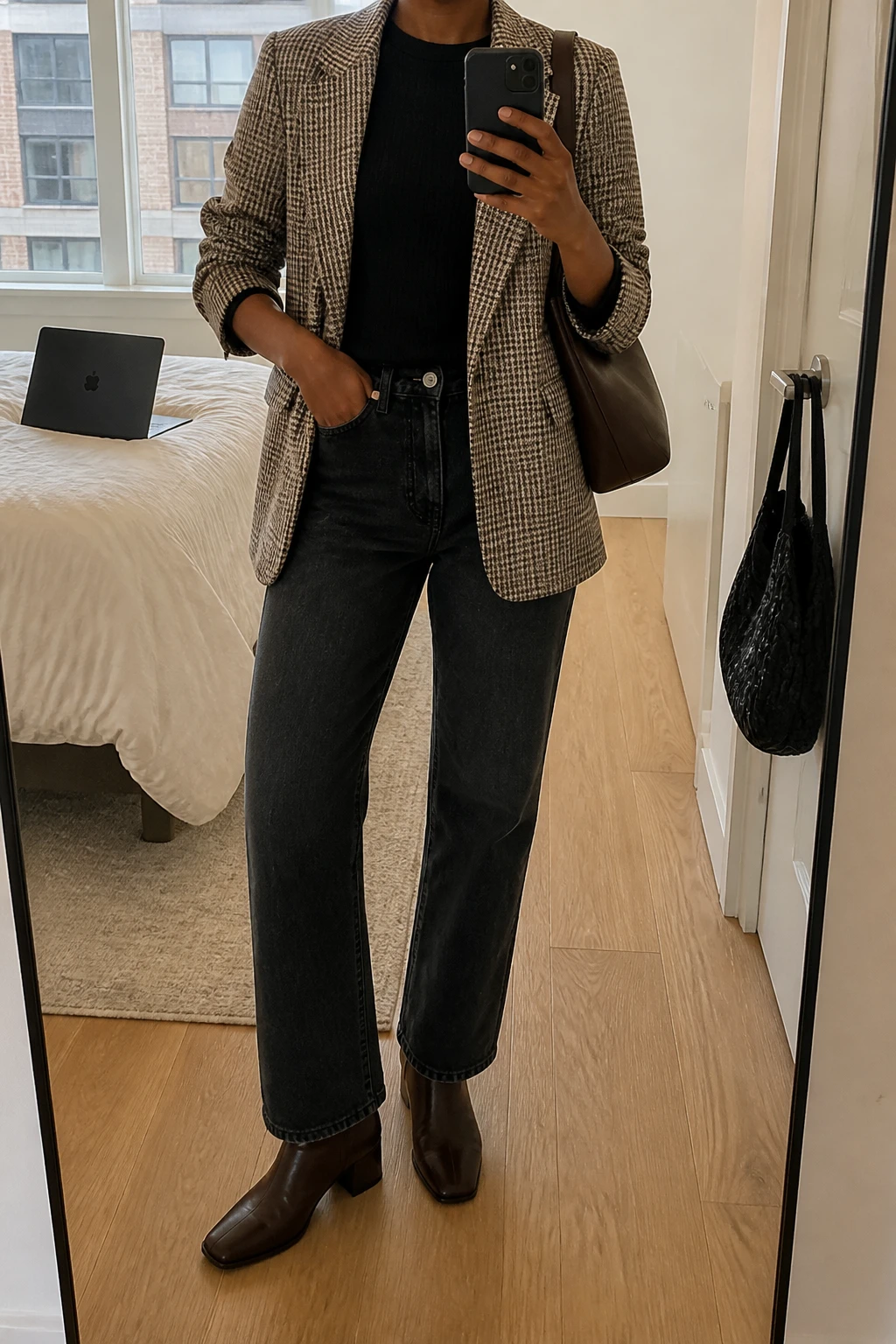 Image Prompt: Mirror selfie of a black woman wearing a brown and beige checked wool-blend blazer with slightly pushed-up sleeves, a black fitted fine-knit crewneck sweater tucked into washed black straight-leg jeans, dark brown leather heeled ankle boots, and a medium chocolate brown leather shoulder bag, holding a phone in front of a mirror, cropped at the NECK so the face does not show, with a relaxed casual selfie pose, in a stylish city apartment, soft natural window light, a laptop on the bed and a handbag hanging on a door handle, subtle denim creases and believable blazer texture, latest iPhone photo quality, no additional clothing or accessories.