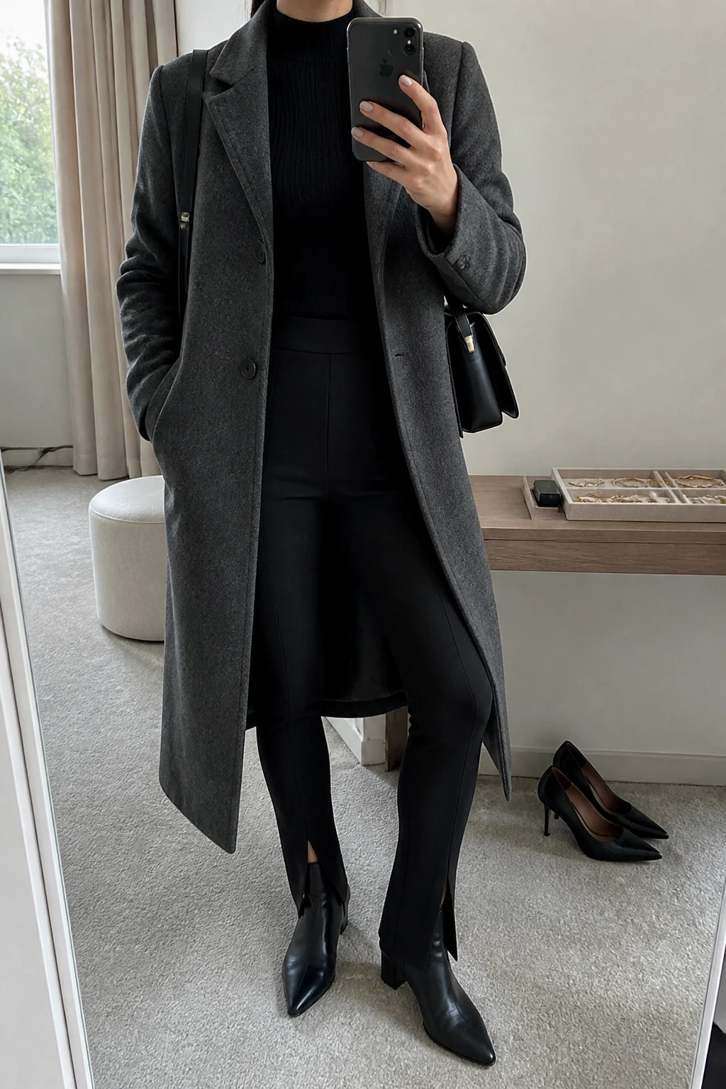 Image Prompt: Mirror selfie of a woman wearing a long charcoal grey tailored wool coat over a black fitted cashmere mock-neck sweater, black high-waisted thick ponte-knit split-hem leggings with small front ankle slits, black leather pointed-toe low block-heel ankle boots, and a structured black leather shoulder bag, holding a phone in front of a mirror, cropped at the NECK so the face does not show, with a relaxed casual selfie pose, in a minimal dressing room, soft natural window light, a jewelry tray on the vanity and a pair of heels on the floor nearby, realistic grey wool coat, natural black ponte fabric and polished casual styling, latest iPhone photo quality, no additional clothing or accessories.