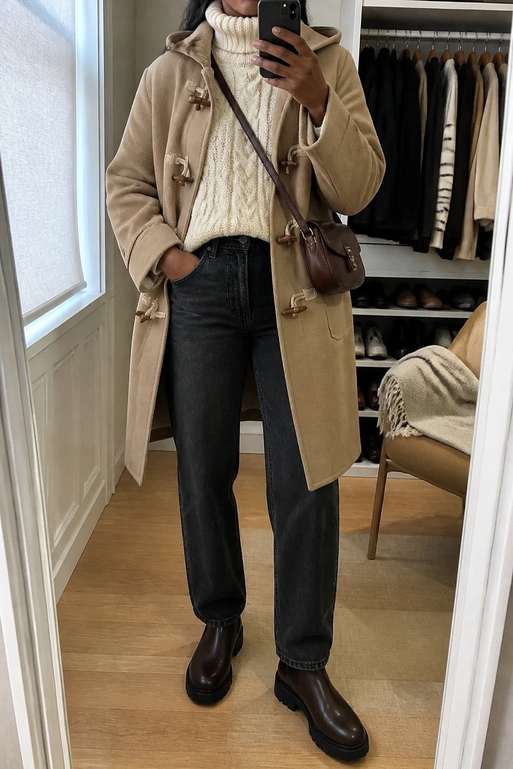 Image Prompt: Mirror selfie of a black woman wearing a camel wool-blend duffle coat with wooden toggle closures over an ivory oversized chunky cable-knit turtleneck sweater, washed black high-waisted straight-leg denim jeans, dark brown leather chunky Chelsea boots with flat rubber soles, and a small chocolate brown leather crossbody bag, holding a phone in front of a mirror, cropped at the NECK so the face does not show, with a relaxed casual selfie pose, in a walk-in closet mirror, soft natural window light, an open closet with clothes visible and a folded blanket on a chair, realistic camel wool fabric, natural ivory chunky knit texture, slightly creased washed black denim, latest iPhone photo quality, no additional clothing or accessories.