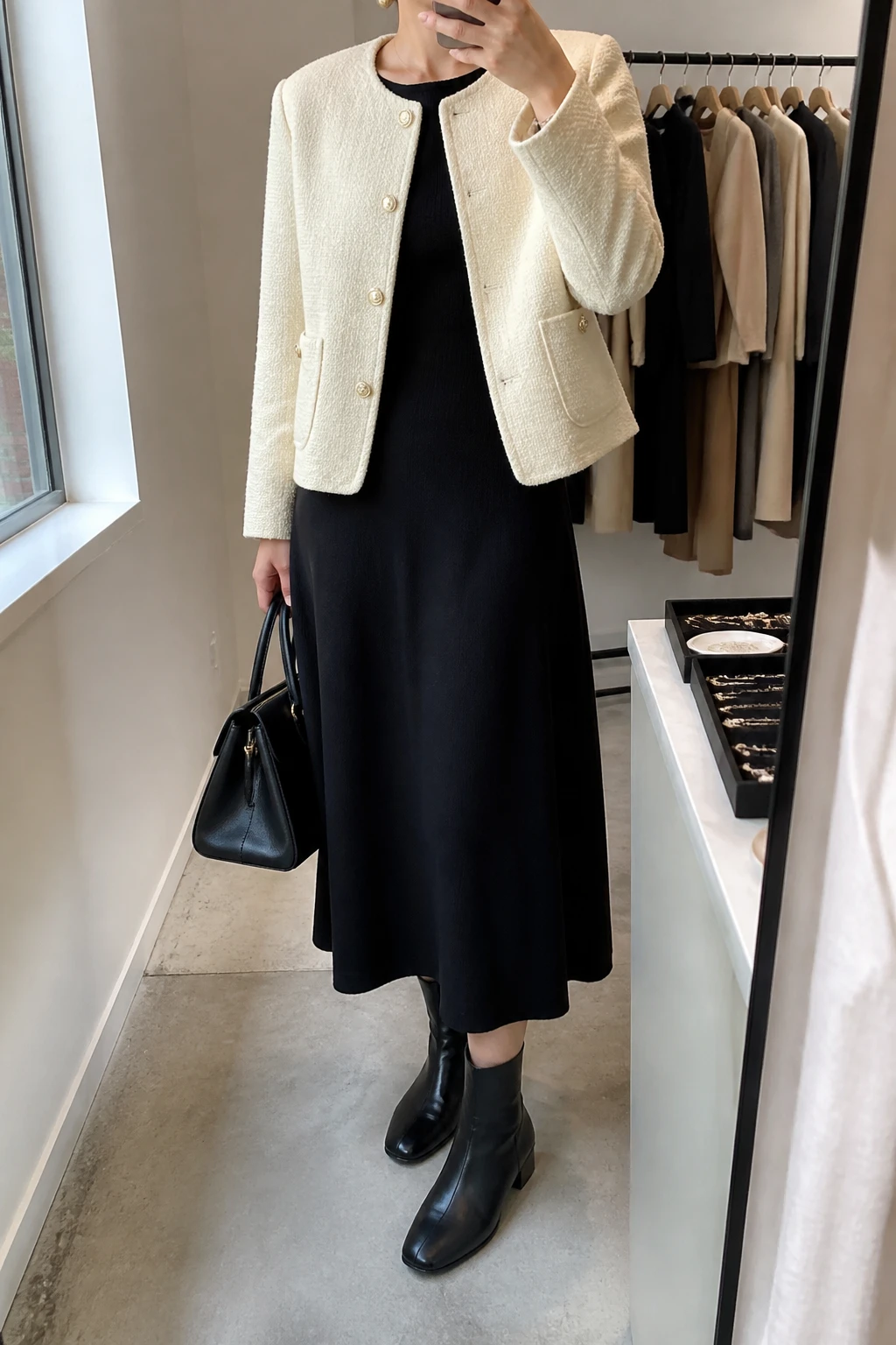 Image Prompt: Mirror selfie of a woman wearing a cream collarless tweed jacket with delicate gold buttons over a black long-sleeve fine-knit midi dress with a modest round neckline and softly fitted waist, black leather low block-heel ankle boots, a structured black leather handbag, and delicate gold stud earrings, holding a phone in front of a mirror, cropped at the NECK so the face does not show, with a relaxed casual selfie pose, in a fashion studio mirror corner, soft natural window light, a clothing rack with garments behind and a jewelry tray on the vanity, realistic cream tweed texture, natural black knit dress folds and elegant everyday proportions, latest iPhone photo quality, no additional clothing or accessories.