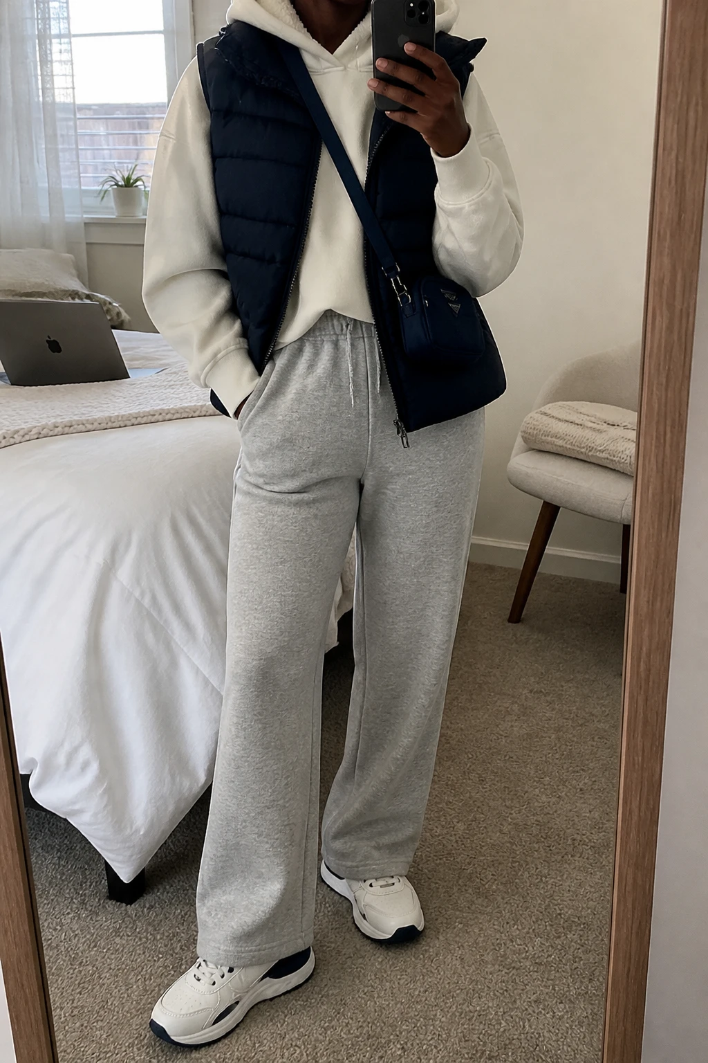 Image Prompt: Mirror selfie of a black woman wearing a navy insulated quilted puffer vest over a cream oversized fleece-lined hoodie, heather grey high-waisted straight-leg knit joggers, white chunky leather sneakers with navy detailing, and a small navy nylon crossbody bag, holding a phone in front of a mirror, cropped at the NECK so the face does not show, with a relaxed casual selfie pose, in a cozy bedroom mirror setup, soft natural window light, a laptop on the bed and a folded blanket on a chair, realistic navy quilted fabric, natural cream hoodie folds and heather grey jogger texture, latest iPhone photo quality, no additional clothing or accessories.