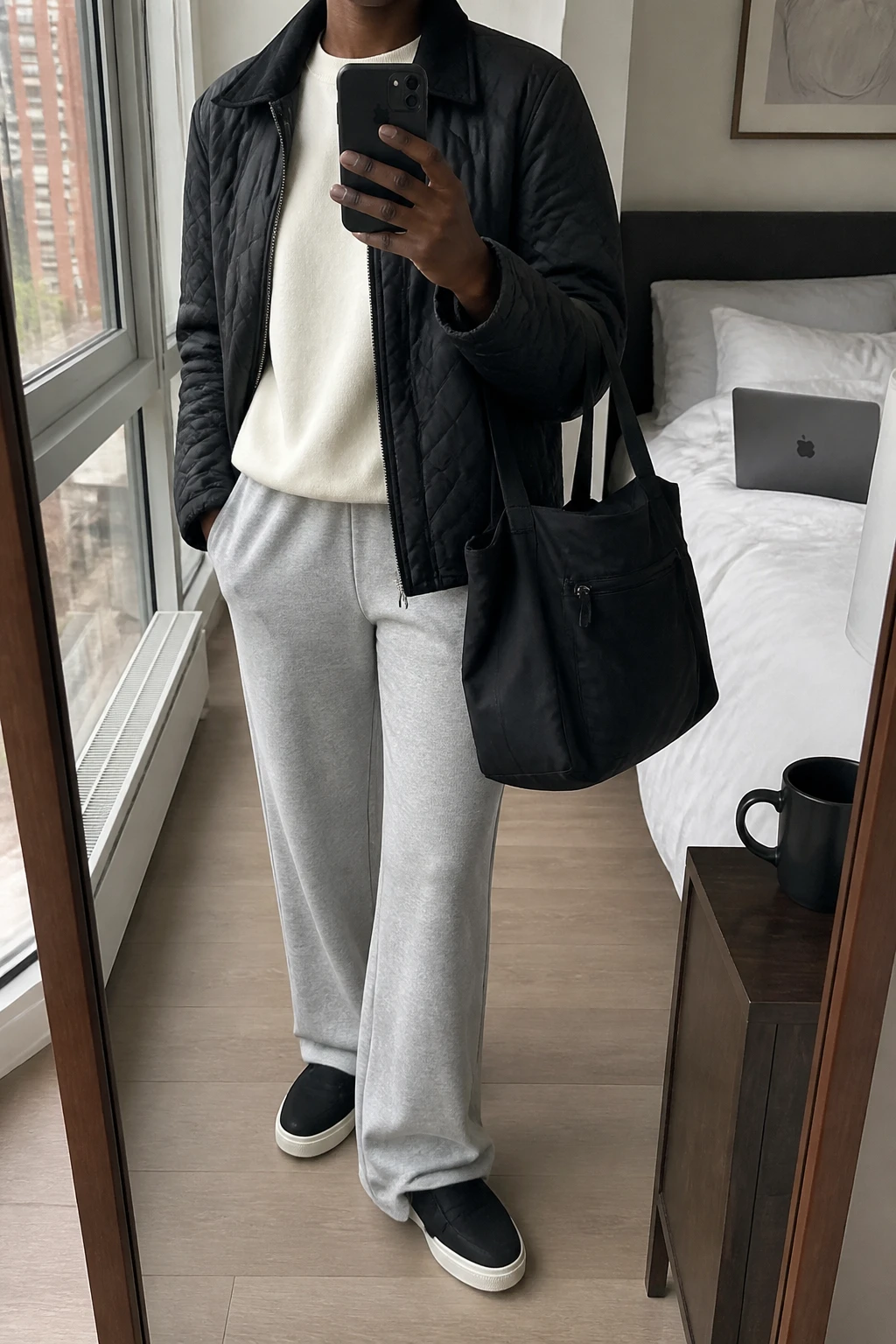 Image Prompt: Mirror selfie of a black woman wearing a black hip-length lightweight quilted insulated jacket over an ivory oversized cotton crewneck sweatshirt, heather grey straight-leg fleece sweatpants with a smooth elastic waistband, black cushioned slip-on sneakers with white rubber soles, and a medium black zippered nylon tote bag, holding a phone in front of a mirror, cropped at the NECK so the face does not show, with a relaxed casual selfie pose, in a stylish city apartment, soft natural window light, a laptop on the bed and a coffee mug on a bedside table, realistic quilted jacket fabric, natural sweatpant folds and casual winter airport styling, latest iPhone photo quality, no additional clothing or accessories.