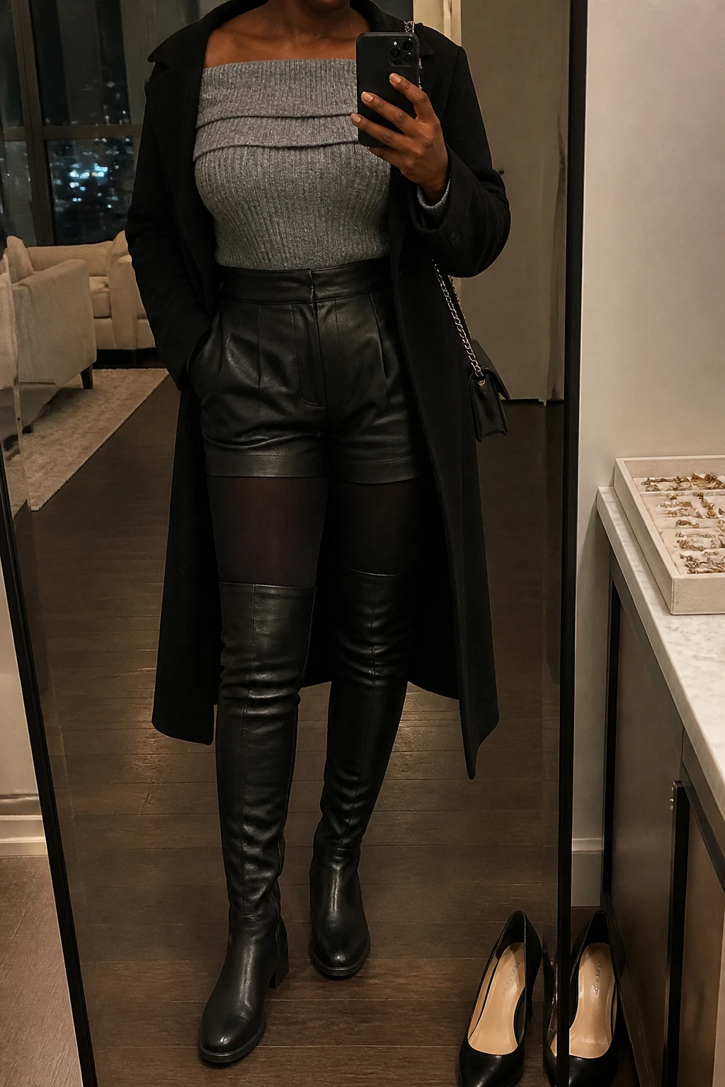 Image Prompt: Mirror selfie of a black woman wearing a heather grey fitted chunky ribbed off-shoulder sweater with a folded neckline tucked into black high-waisted faux-leather tailored mini shorts, black opaque fleece-lined thermal tights, black leather over-the-knee boots with low block heels, a long black tailored wool coat worn open, and a small silver chain-strap shoulder bag, holding a phone in front of a mirror, cropped at the NECK so the face does not show, with a relaxed casual selfie pose, in a stylish city apartment, warm moody lighting, a jewelry tray on the vanity and a pair of heels on the floor, realistic grey chunky-knit texture, natural black faux-leather grain and warm thermal tights, latest iPhone photo quality, no additional clothing or accessories.
