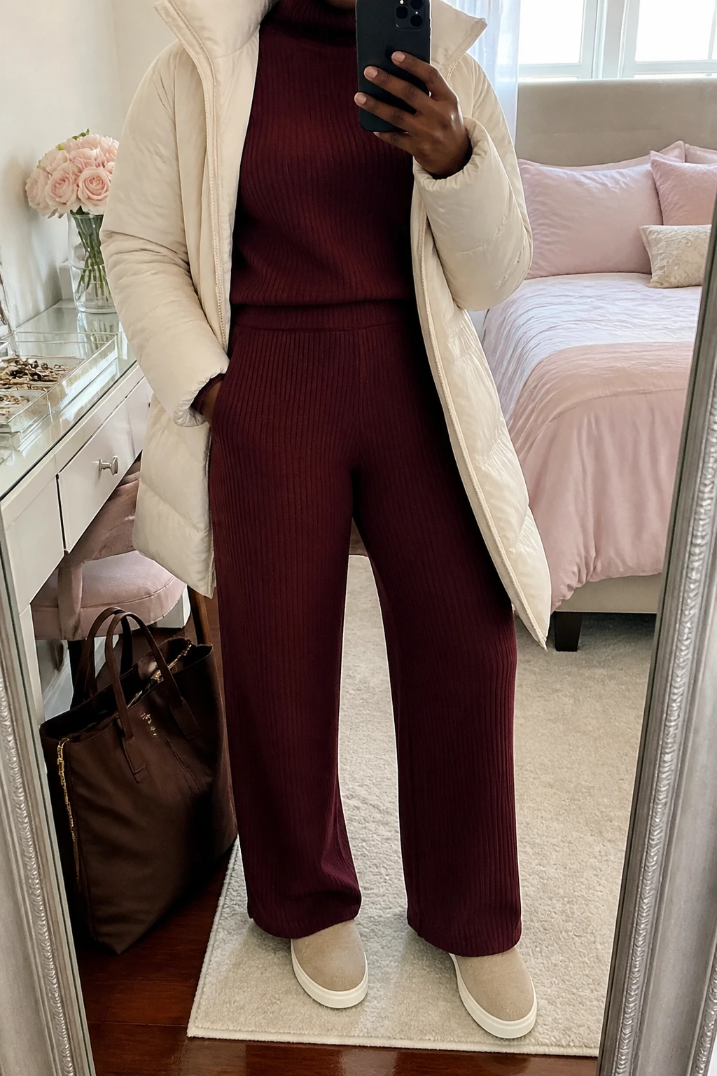 Image Prompt: Mirror selfie of a black woman wearing a matching burgundy ribbed knit travel set consisting of a relaxed long-sleeve mock-neck sweater and high-waisted wide-leg trousers with a soft elastic waistband, a cream lightweight insulated puffer coat worn open, beige cushioned slip-on sneakers, and a medium chocolate brown nylon zippered tote bag, holding a phone in front of a mirror, cropped at the NECK so the face does not show, with a relaxed casual selfie pose, in a feminine-themed bedroom, soft natural window light, a tote bag resting on the floor and a jewelry tray on the vanity, realistic burgundy knit texture, natural cream puffer volume and comfortable travel proportions, latest iPhone photo quality, no additional clothing or accessories.