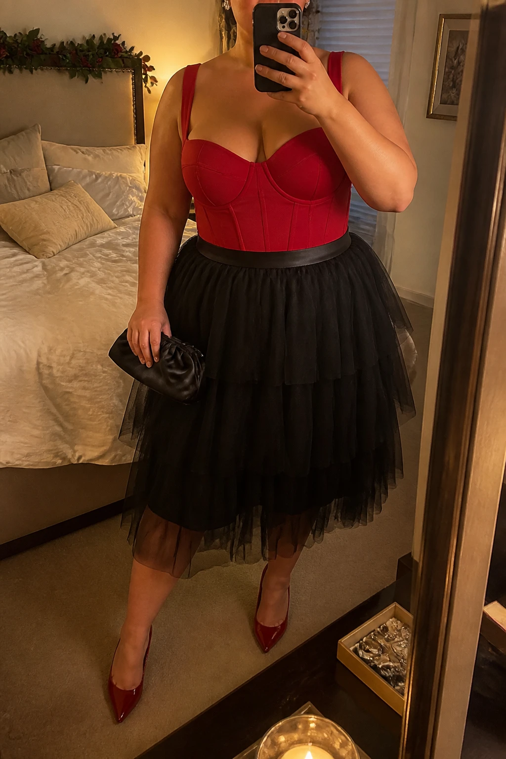 Mirror selfie of a plus size woman wearing a layered black tulle midi skirt with an opaque black lining, a structured red corset-style bodysuit with wide supportive straps, red pointed pumps, a small black satin clutch, and crystal stud earrings, clearly showing the exact clothing items, black tulle, red structured top, footwear, and accessories listed, holding a phone in front of a mirror, cropped at the neck so her face is hidden, relaxed dramatic selfie pose, in a cozy bedroom mirror setup with evening golden-hour lighting, soft pillows on an unmade bed and a jewelry tray on the vanity, realistic formal birthday dinner appearance, latest iPhone photo quality.