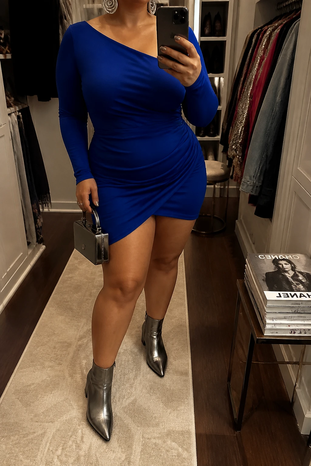 Mirror selfie of a plus size woman wearing a sapphire blue long-sleeve mini dress with an asymmetric neckline, gentle waist ruching, and a softly fitted silhouette, metallic gunmetal pointed ankle boots with low block heels, a small gunmetal box bag, and silver spiral earrings, clearly showing the exact clothing items, sapphire color, asymmetric neckline, ruching, footwear, and accessories listed, holding a phone in front of a mirror, cropped at the neck so her face does not appear, relaxed bold selfie pose, in a walk-in closet mirror with warm moody lighting, an open closet with clothes visible and a stack of fashion magazines on a side table, realistic unforgettable birthday night appearance, latest iPhone photo quality.