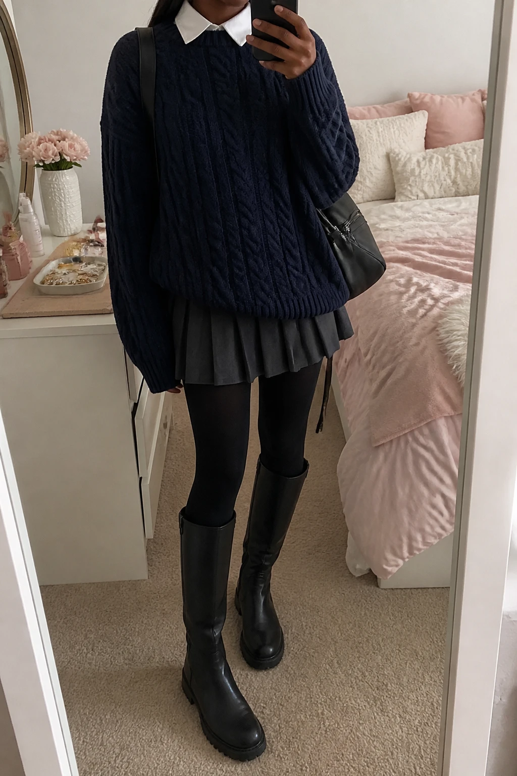 Image Prompt: Mirror selfie of a black woman wearing an oversized navy chunky cable-knit crewneck sweater layered over a white cotton collared shirt with the collar visible, a charcoal grey wool-blend pleated mini skirt, black opaque fleece-lined tights, black leather knee-high flat boots with rubber soles, and a small black nylon backpack worn over one shoulder, holding a phone in front of a mirror, cropped at the NECK so the face does not show, with a relaxed casual selfie pose, in a feminine-themed bedroom, soft natural window light, a jewelry tray on the vanity and soft pillows on an unmade bed, realistic navy cable-knit texture, natural pleated skirt movement and believable black tights, latest iPhone photo quality, no additional clothing or accessories.