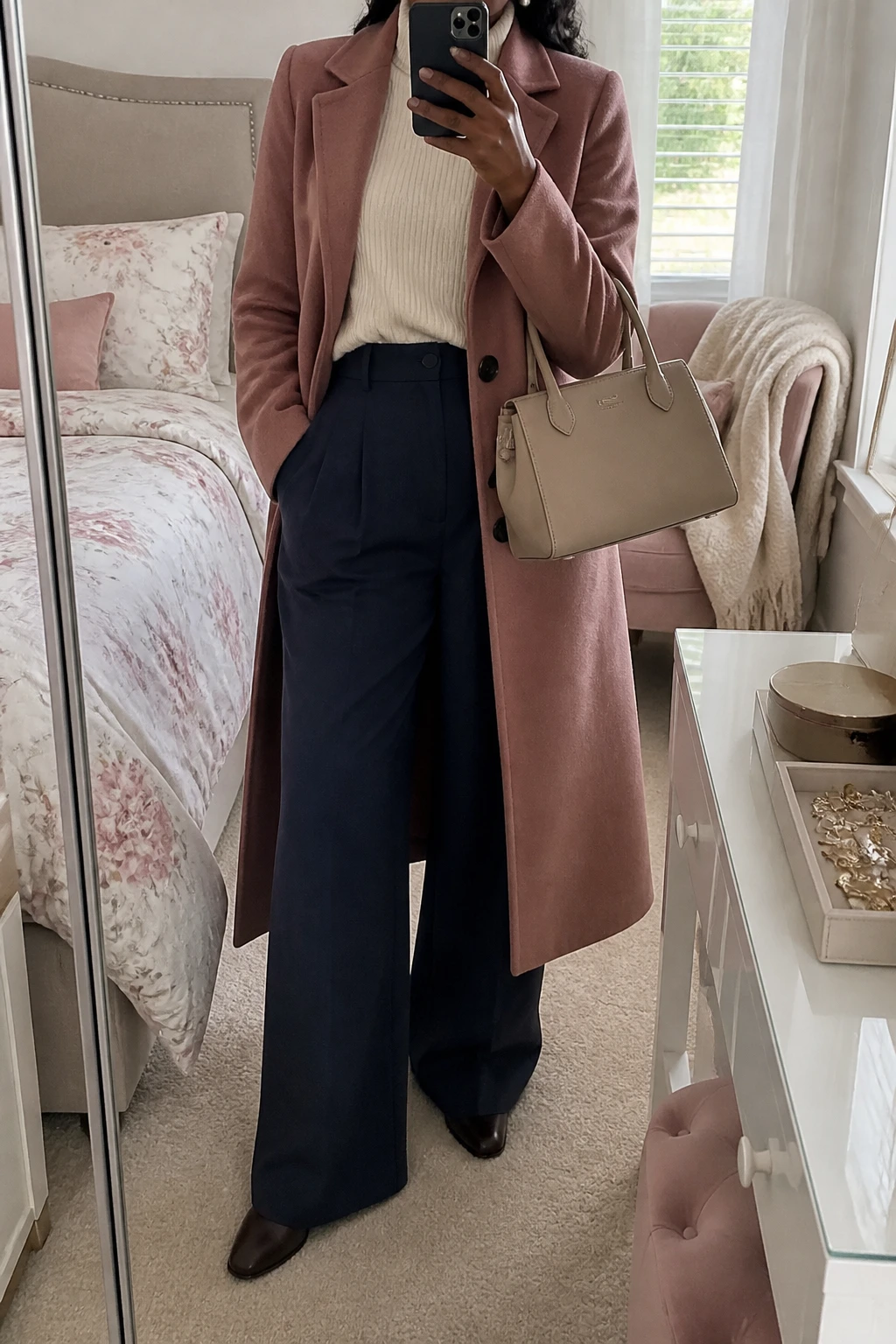 Image Prompt: Mirror selfie of a black woman wearing a long dusty rose tailored wool coat over a cream relaxed-fit ribbed turtleneck sweater, navy high-waisted wide-leg wool trousers with flowing legs and pressed creases, dark brown leather flat ankle boots, a structured beige leather handbag, and delicate pearl stud earrings, holding a phone in front of a full-length mirror, cropped at the NECK so the face does not show, with a relaxed casual selfie pose, in a feminine-themed bedroom, soft natural window light, a jewelry tray on the vanity and a folded blanket on a chair, realistic dusty rose wool texture, natural navy trouser folds and soft cream knit fabric, true-to-life muted colors, latest iPhone photo quality, no additional clothing or accessories.