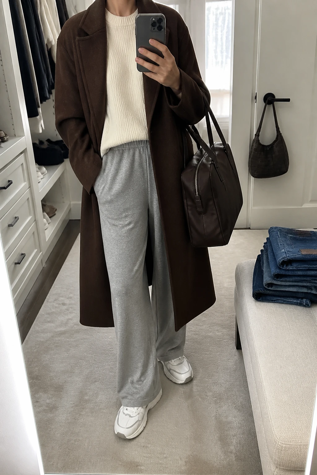Image Prompt: Mirror selfie of a woman wearing a long chocolate brown lightweight wool-blend coat over a cream oversized fine-knit crewneck sweater, heather grey relaxed wide-leg jersey lounge trousers with an elastic waistband, white cushioned leather sneakers with grey details, and a medium dark brown nylon zippered travel tote bag, holding a phone in front of a mirror, cropped at the NECK so the face does not show, with a relaxed casual selfie pose, in a walk-in closet mirror, soft natural window light, folded denim jeans on a bench and a handbag hanging on a door handle, realistic chocolate wool texture, natural grey jersey folds and polished casual airport styling, latest iPhone photo quality, no additional clothing or accessories.