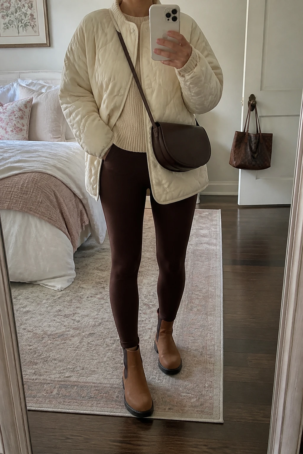 Image Prompt: Mirror selfie of a woman wearing a cream oversized quilted insulated hip-length jacket over an oatmeal chunky ribbed crewneck sweater, chocolate brown high-waisted fleece-lined leggings, tan leather flat Chelsea boots with rubber soles, and a small dark brown leather crossbody bag, holding a phone in front of a mirror, cropped at the NECK so the face does not show, with a relaxed casual selfie pose, in a feminine-themed bedroom, soft natural window light, soft pillows on an unmade bed and a handbag hanging on a door handle, realistic cream quilted fabric, warm brown leggings and natural knit texture, latest iPhone photo quality, no additional clothing or accessories.