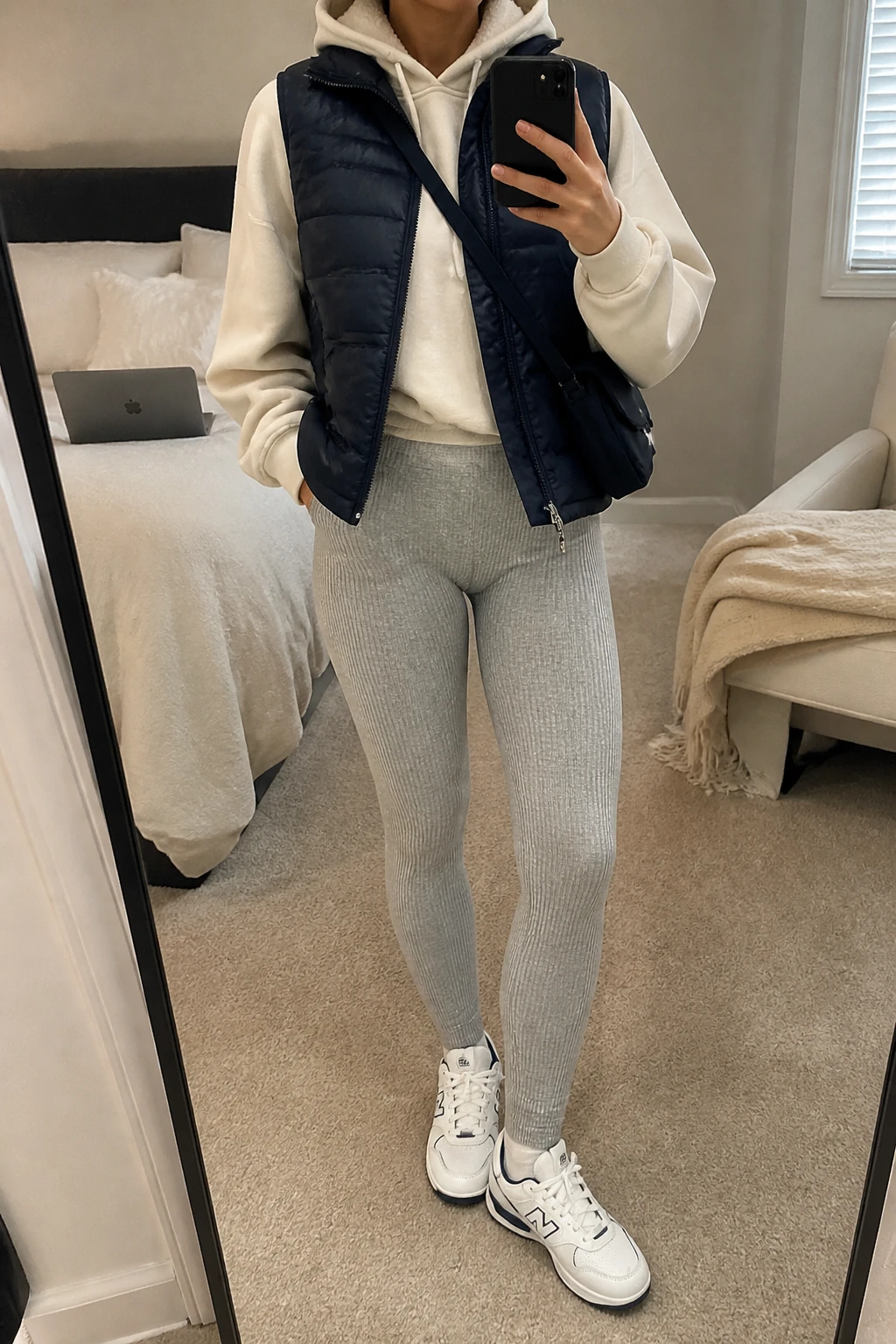 Image Prompt: Mirror selfie of a woman wearing a navy insulated quilted puffer vest over a cream oversized fleece-lined hoodie, heather grey high-waisted thick ribbed leggings, white chunky leather sneakers with navy detailing, and a small navy nylon crossbody bag, holding a phone in front of a mirror, cropped at the NECK so the face does not show, with a relaxed casual selfie pose, in a cozy bedroom mirror setup, soft indoor lighting, a laptop on the bed and a folded blanket on a chair, realistic navy quilted fabric, natural grey ribbed leggings texture and relaxed sporty styling, latest iPhone photo quality, no additional clothing or accessories.