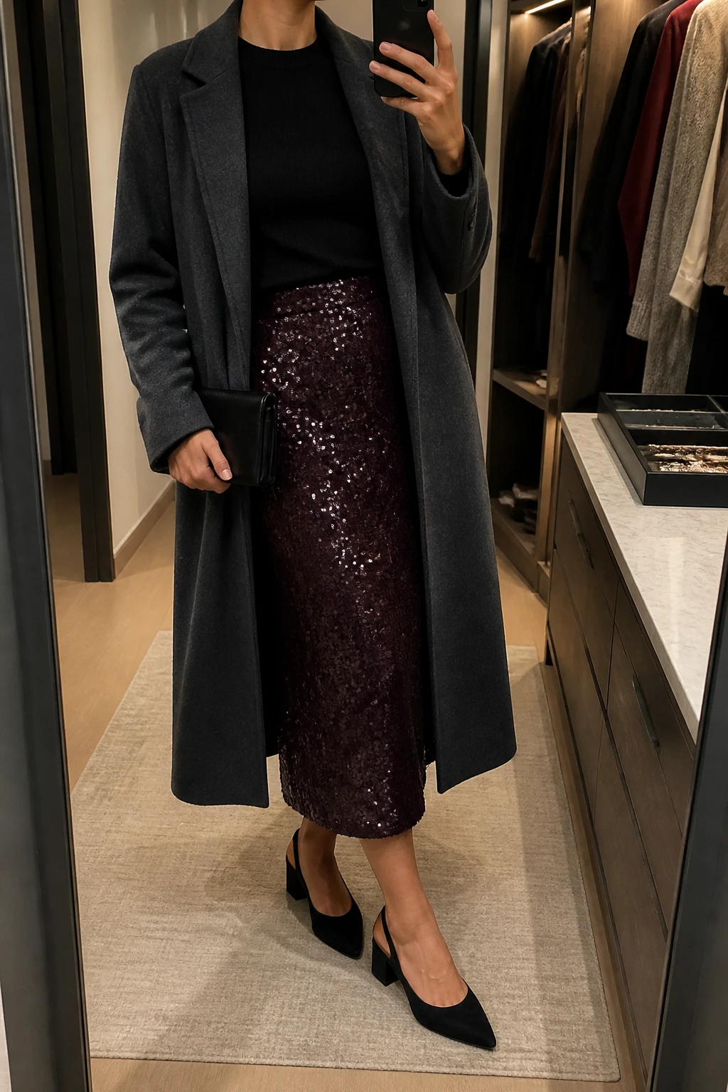 Image Prompt: Mirror selfie of a woman wearing a black fitted cashmere crewneck sweater tucked into a deep plum sequin midi skirt, black suede pointed-toe low block heels, a long charcoal grey tailored wool coat worn open, a small black satin clutch, and delicate silver stud earrings, holding a phone in front of a mirror, cropped at the NECK so the face does not show, with a relaxed casual selfie pose, in a walk-in closet mirror, warm moody indoor lighting, an open closet with clothes visible and a jewelry tray on the vanity, realistic deep plum sequins with subtle natural reflections, soft black cashmere texture, latest iPhone photo quality, no additional clothing or accessories.