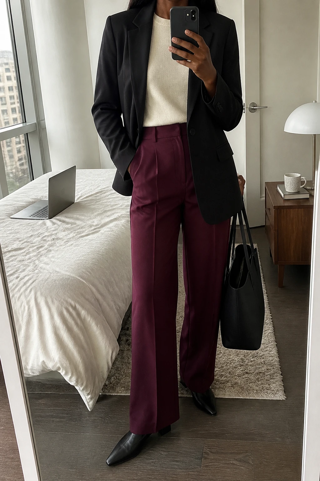 Image Prompt: Mirror selfie of a black woman wearing a black tailored single-breasted blazer over a cream fine-knit crewneck sweater, burgundy high-waisted wide-leg tailored trousers, black leather pointed-toe low block heels, and a structured black leather tote bag, holding a phone in front of a mirror, cropped at the NECK so the face does not show, with a relaxed casual selfie pose, in a stylish city apartment, soft natural window light, a laptop on the bed and a coffee mug on a bedside table, realistic burgundy trouser fabric, natural professional styling, latest iPhone photo quality, no additional clothing or accessories.