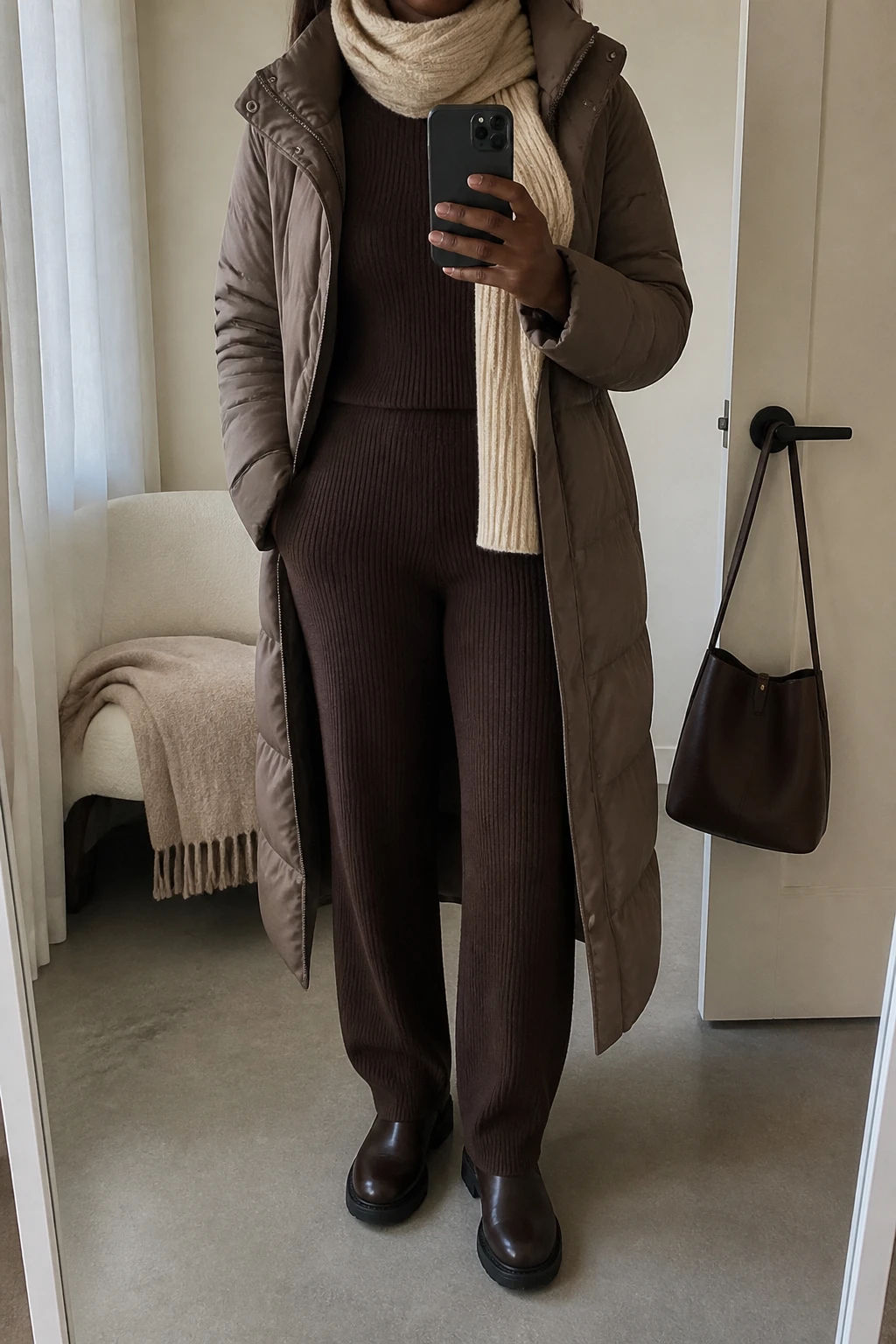 Image Prompt: Mirror selfie of a black woman wearing a long taupe insulated quilted winter coat left open over a chocolate brown fitted ribbed knit turtleneck sweater and matching chocolate brown straight-leg ribbed knit trousers, dark brown leather flat ankle boots with thick rubber soles, and a cream ribbed wool scarf wrapped loosely around the neck, holding a phone in front of a mirror, cropped at the NECK so the face does not show, with a relaxed casual selfie pose, in a minimal dressing room, soft natural window light, a folded blanket on a chair and a handbag hanging on a door handle, natural quilted fabric, realistic matching knit texture and warm neutral colors, latest iPhone photo quality, no additional clothing or accessories.