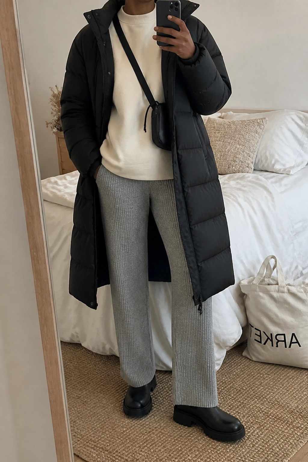 Image Prompt: Mirror selfie of a black woman wearing a long black insulated quilted down puffer coat over a cream thick fleece-lined crewneck sweatshirt, heather grey straight-leg ribbed knit trousers, black waterproof leather chunky Chelsea boots with flat lug soles, and a small black nylon crossbody bag, holding a phone in front of a mirror, cropped at the NECK so the face does not show, with a relaxed casual selfie pose, in a cozy bedroom mirror setup, soft indoor lighting, soft pillows on an unmade bed and a tote bag resting on the floor, realistic down coat volume, natural fleece and knit textures, latest iPhone photo quality, no additional clothing or accessories.