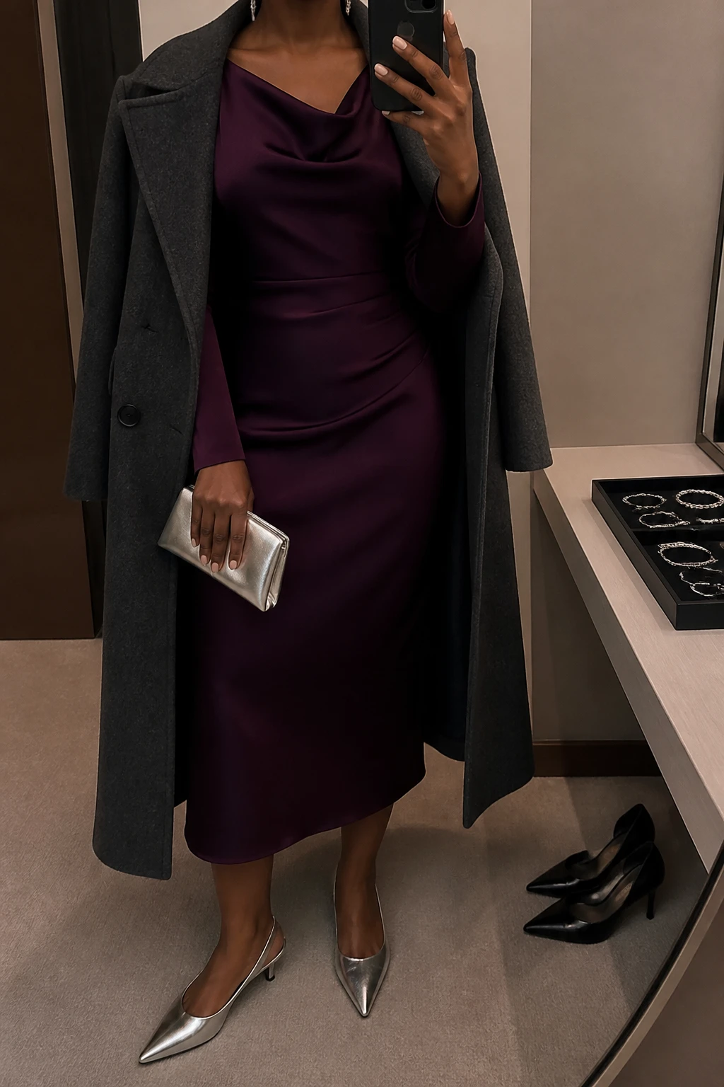 Image Prompt: Mirror selfie of a black woman wearing a deep plum long-sleeve satin midi dress with a draped neckline and softly fitted waist, silver metallic pointed-toe kitten heels, a small silver satin evening clutch, delicate silver drop earrings, and a long charcoal grey wool coat worn open, holding a phone in front of a mirror, cropped at the NECK so the face does not show, with a relaxed casual selfie pose, in a minimal dressing room, warm moody lighting, a jewelry tray on the vanity and a pair of heels on the floor nearby, realistic satin draping, subdued rich plum tones and natural proportions, latest iPhone photo quality, no additional clothing or accessories.