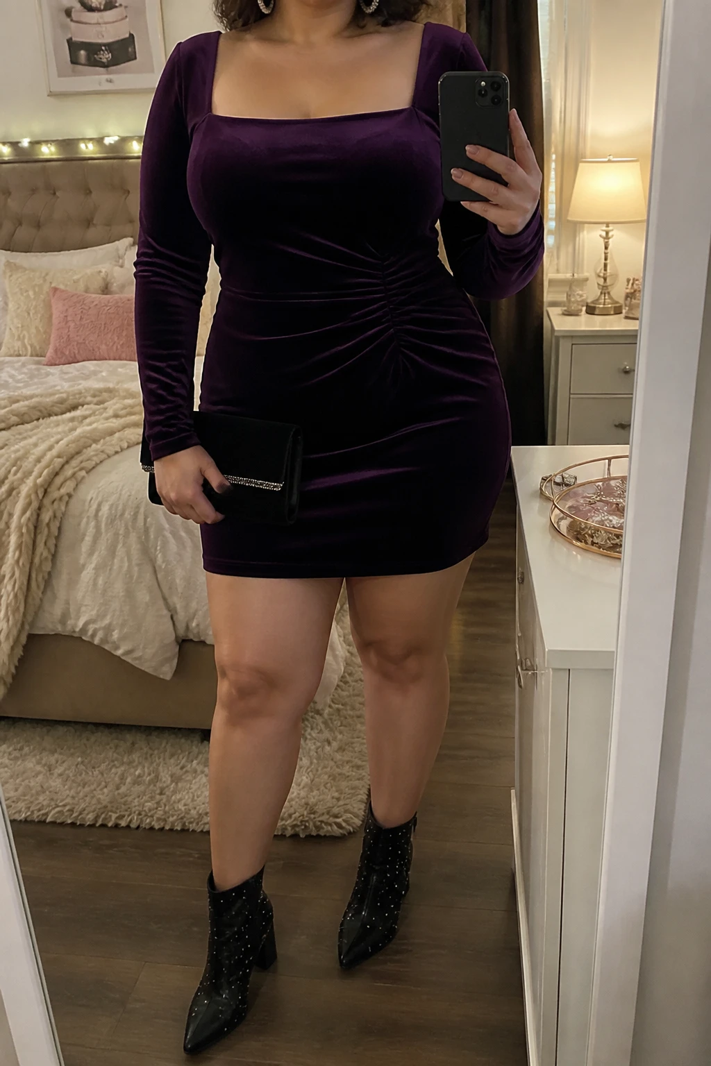 Mirror selfie of a plus size woman wearing a plum purple velvet mini dress with long sleeves, a square neckline, and soft gathering at the waist, black pointed ankle boots with small crystal details and block heels, a black velvet clutch, and silver crystal hoop earrings, clearly showing the exact clothing items, plum velvet, waist gathering, footwear, and accessories listed, holding a phone in front of a mirror, cropped at the neck so the face does not appear, relaxed elegant selfie stance, in a feminine-themed bedroom with warm moody lighting, soft pillows on an unmade bed and a jewelry tray on the vanity, realistic winter birthday outfit, latest iPhone photo quality.