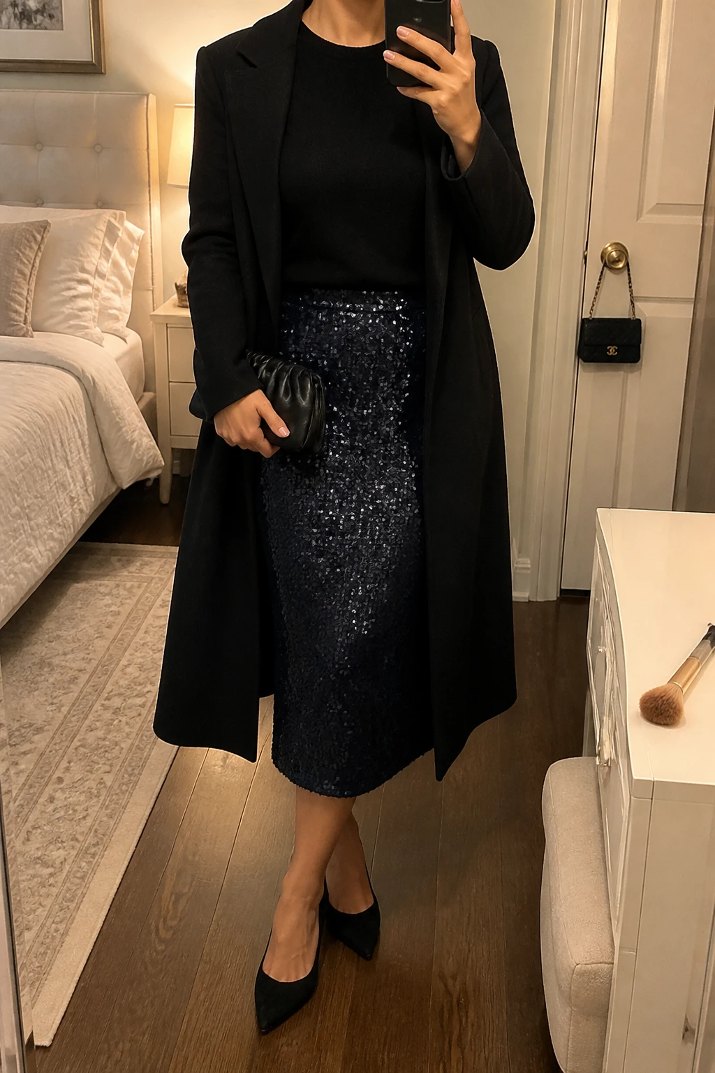 Image Prompt: Mirror selfie of a woman wearing a black fitted cashmere crewneck sweater tucked into a midnight navy sequin midi skirt, black suede pointed-toe kitten heels, a black satin mini clutch, delicate silver stud earrings, and a long black tailored wool coat worn open, holding a phone in front of a mirror, cropped at the NECK so the face does not show, with a relaxed casual selfie pose, in a feminine-themed bedroom, warm moody indoor lighting, a makeup brush on the vanity and a small handbag hanging on a door handle, natural sequin reflections, realistic soft sweater texture, latest iPhone photo quality, no additional clothing or accessories.