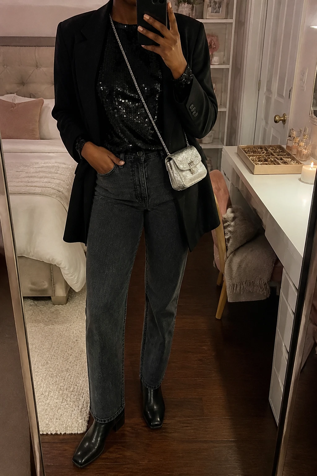 Image Prompt: Mirror selfie of a black woman wearing a black long-sleeve fitted sequin top with a simple crew neckline, dark charcoal grey straight-leg denim jeans, a black oversized wool-blend blazer worn open, black leather low block-heel ankle boots, and a small silver metallic crossbody bag, holding a phone in front of a mirror, cropped at the NECK so the face does not show, with a relaxed casual selfie pose, in a feminine-themed bedroom, warm moody indoor lighting, a jewelry tray on the vanity and a folded blanket on a chair, realistic black sequin reflections without exaggerated sparkle, natural denim texture and casual mirror selfie framing, latest iPhone photo quality, no additional clothing or accessories.
