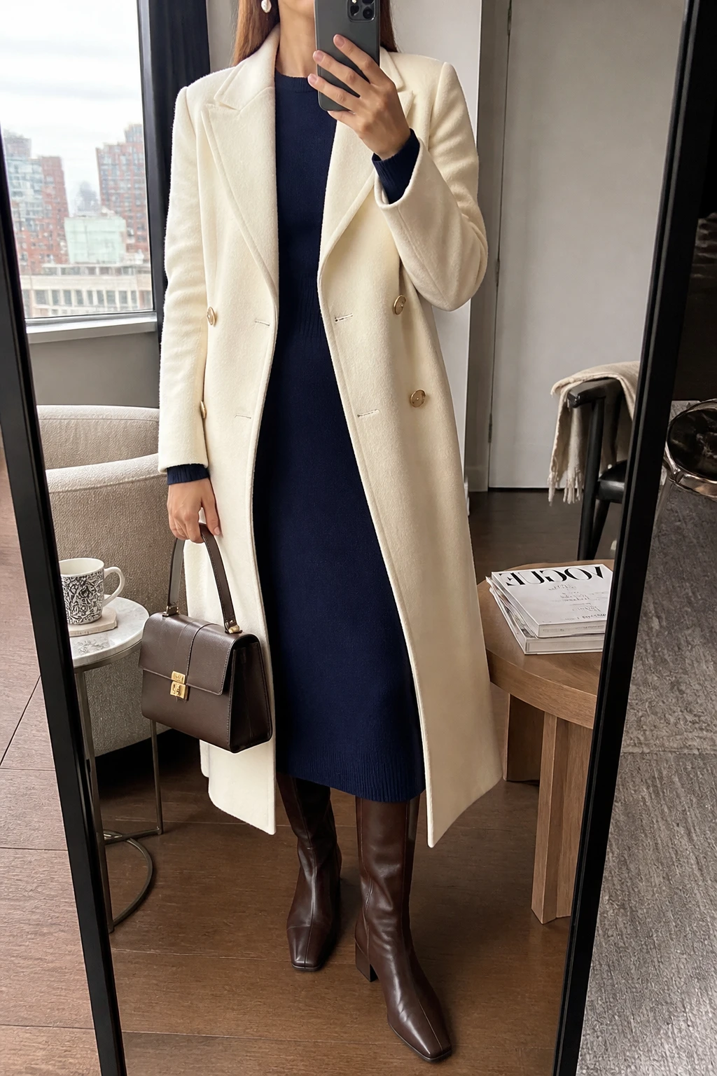 Image Prompt: Mirror selfie of a woman wearing a long cream double-breasted tailored wool coat over a navy long-sleeve fine-knit cashmere midi dress with a modest round neckline and gently defined waist, chocolate brown polished leather knee-high boots with low block heels, a structured dark brown leather top-handle handbag, and classic pearl drop earrings, holding a phone in front of a mirror, cropped at the NECK so the face does not show, with a relaxed casual selfie pose, in a stylish city apartment, soft natural window light, a coffee mug on a side table and a stack of fashion magazines on a side table, realistic cream wool texture, natural navy knit dress folds and understated classic styling, latest iPhone photo quality, no additional clothing or accessories.