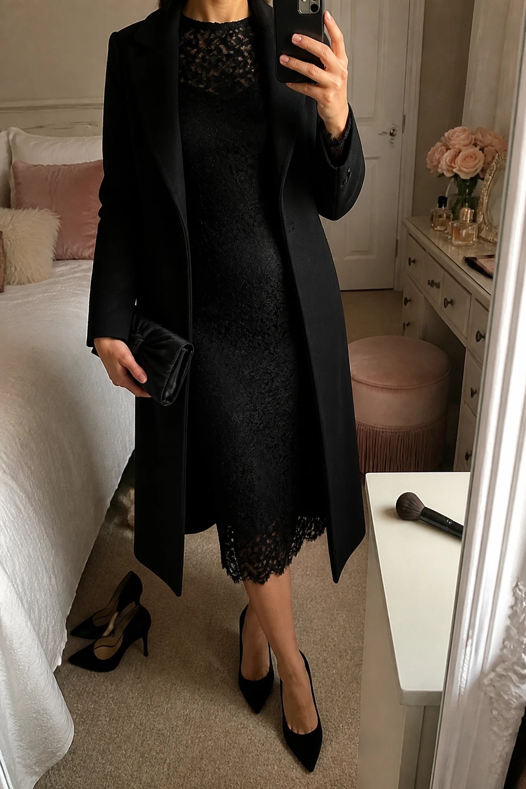 Image Prompt: Mirror selfie of a woman wearing a black long-sleeve lace midi dress with opaque black lining, a fitted waist and delicate scalloped sleeve edges, black velvet pointed-toe heels, a small black satin evening clutch, pearl stud earrings, and a long black tailored wool coat worn open, holding a phone in front of a mirror, cropped at the NECK so the face does not show, with a relaxed casual selfie pose, in a feminine-themed bedroom, soft warm indoor lighting, a makeup brush on the counter and a pair of heels on the floor nearby, realistic lace detailing, soft fabric texture and natural pose, latest iPhone photo quality, no additional clothing or accessories.