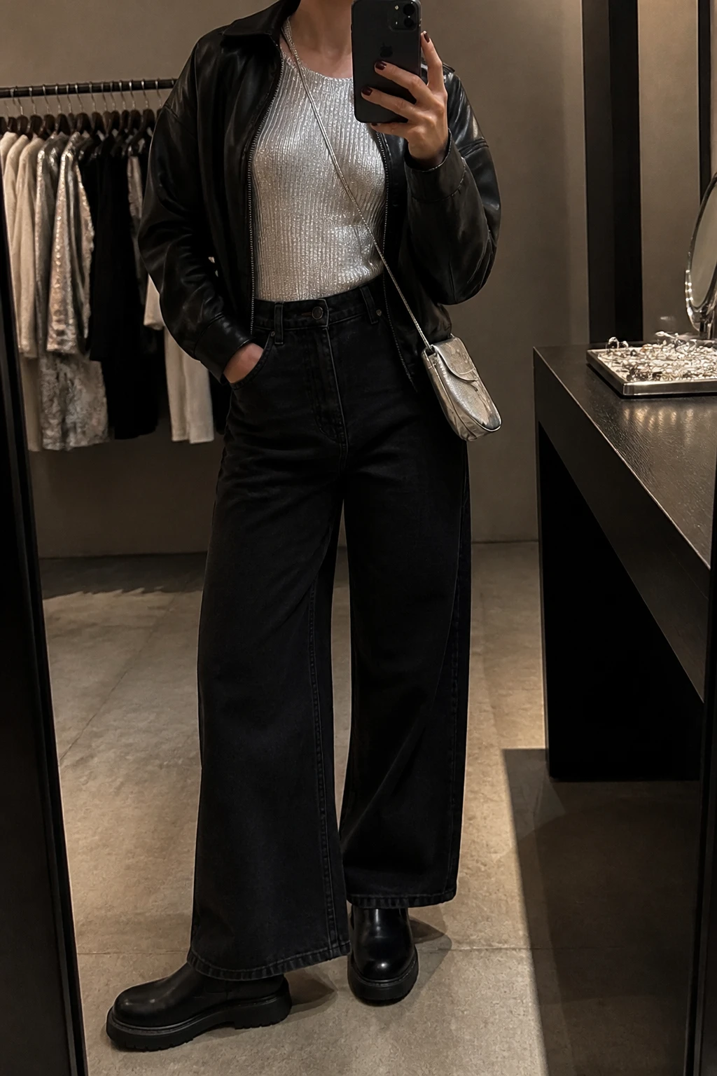 Image Prompt: Mirror selfie of a woman wearing a silver metallic long-sleeve fitted knit top, black high-waisted wide-leg denim jeans, a cropped black faux-leather jacket worn open, black leather chunky Chelsea boots with flat soles, and a small metallic silver crossbody bag, holding a phone in front of a mirror, cropped at the NECK so the face does not show, with a relaxed casual selfie pose, in a fashion studio mirror corner, warm moody lighting, a jewelry tray on the vanity and a clothing rack with garments behind, natural silver fabric reflections, realistic denim texture, slightly imperfect casual pose, latest iPhone photo quality, no additional clothing or accessories.