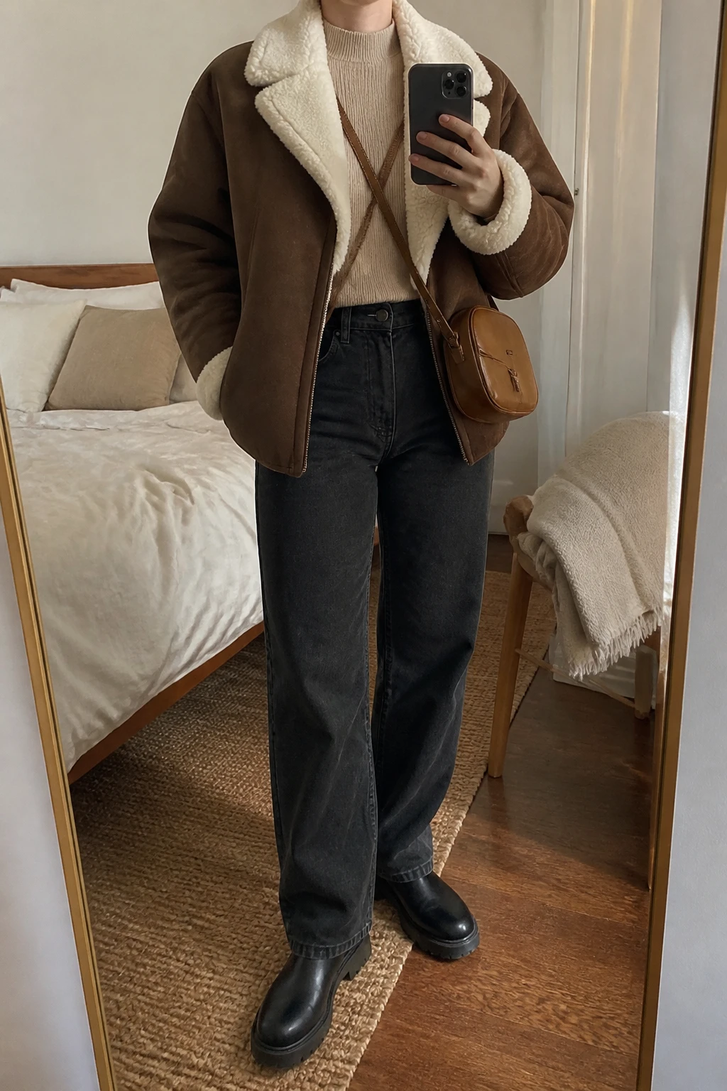 Image Prompt: Mirror selfie of a woman wearing an oversized chocolate brown faux-suede shearling-lined jacket with a thick ivory fleece collar over an oatmeal fitted ribbed mock-neck sweater, washed black high-waisted wide-leg jeans with relaxed full-length legs, black leather chunky Chelsea boots with flat rubber soles, and a small caramel leather crossbody bag, holding a phone in front of a mirror, cropped at the NECK so the face does not show, with a relaxed casual selfie pose, in a cozy bedroom mirror setup, soft indoor lighting, soft pillows on an unmade bed and a folded blanket on a chair, realistic brown faux-suede grain, natural ivory shearling texture, oatmeal ribbed fabric, slightly creased black denim, latest iPhone photo quality, no additional clothing or accessories.