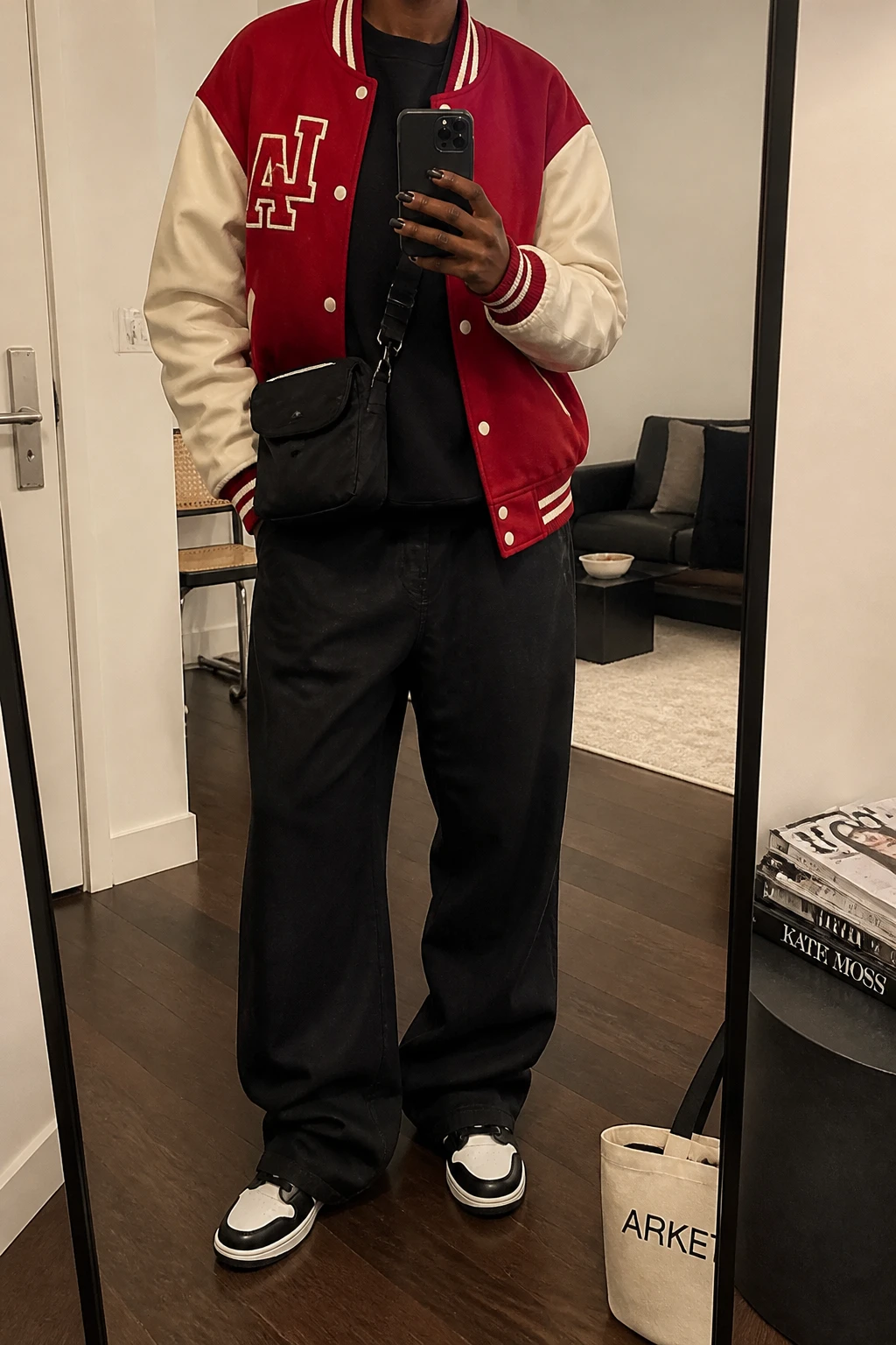 Image Prompt: Mirror selfie of a black woman wearing a cherry red wool varsity jacket with cream faux-leather sleeves over a black heavyweight fleece-lined crewneck sweatshirt, black baggy wide-leg cotton twill trousers, black and white chunky leather sneakers, and a small black nylon crossbody bag, holding a phone in front of a mirror, cropped at the NECK so the face does not show, with a relaxed casual selfie pose, in a stylish city apartment, soft indoor lighting, a stack of fashion magazines on a side table and a tote bag resting on the floor, natural varsity wool texture, realistic red color and relaxed trouser folds, latest iPhone photo quality, no additional clothing or accessories.