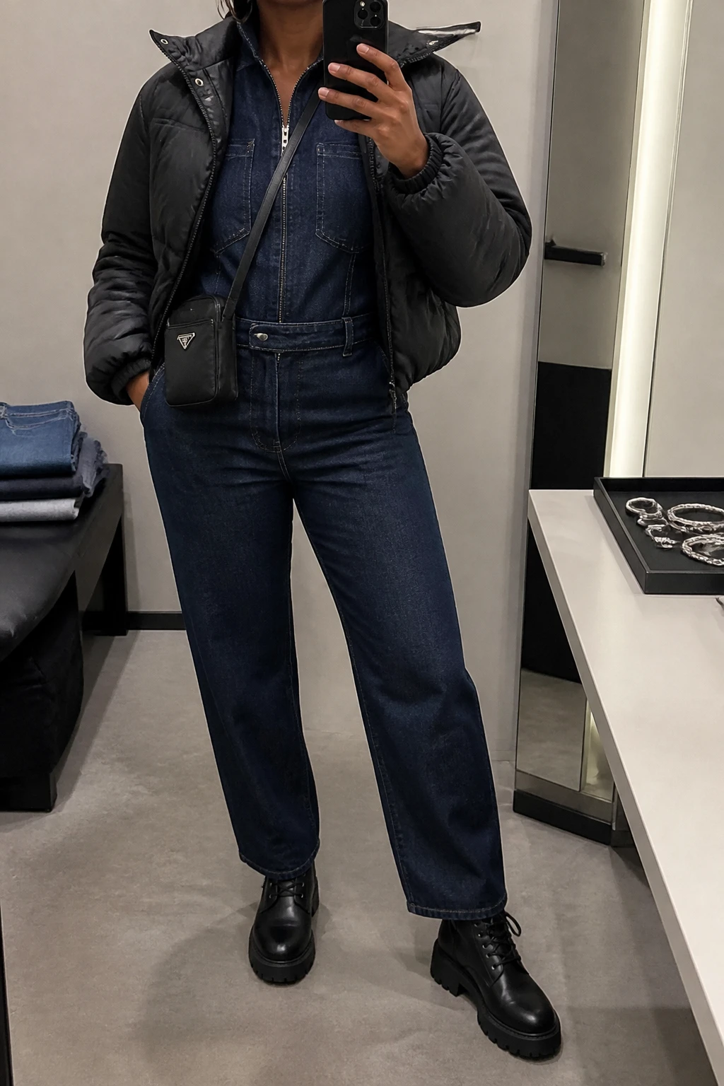 Image Prompt: Mirror selfie of a black woman wearing a dark indigo long-sleeve utility denim jumpsuit with a zip front, defined waist and straight legs, black leather chunky lace-up combat boots, a cropped black quilted winter jacket worn open, a small black nylon crossbody bag, and silver hoop earrings, holding a phone in front of a mirror, cropped at the NECK so the face does not show, with a relaxed casual selfie pose, in a minimal dressing room, soft indoor lighting, folded denim jeans on a bench and a jewelry tray on the vanity, realistic denim texture and natural utility jumpsuit folds, latest iPhone photo quality, no additional clothing or accessories.