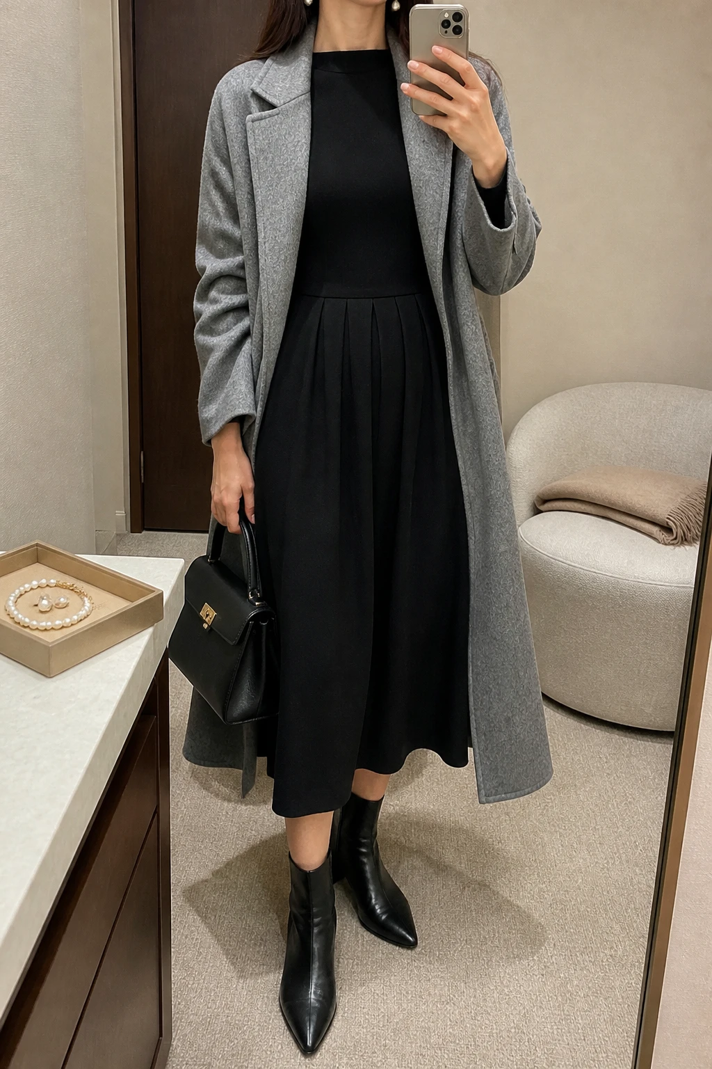 Image Prompt: Mirror selfie of a woman wearing a black long-sleeve wool-blend midi dress with a modest boat neckline and softly pleated skirt, black leather pointed-toe low block-heel ankle boots, a long heather grey wool coat worn open, a structured black leather top-handle bag, and classic white pearl drop earrings, holding a phone in front of a mirror, cropped at the NECK so the face does not show, with a relaxed casual selfie pose, in a minimal dressing room, soft indoor lighting, a jewelry tray on the vanity and a folded blanket on a chair, realistic black wool dress fabric, natural pleated skirt folds and understated elegant styling, latest iPhone photo quality, no additional clothing or accessories.