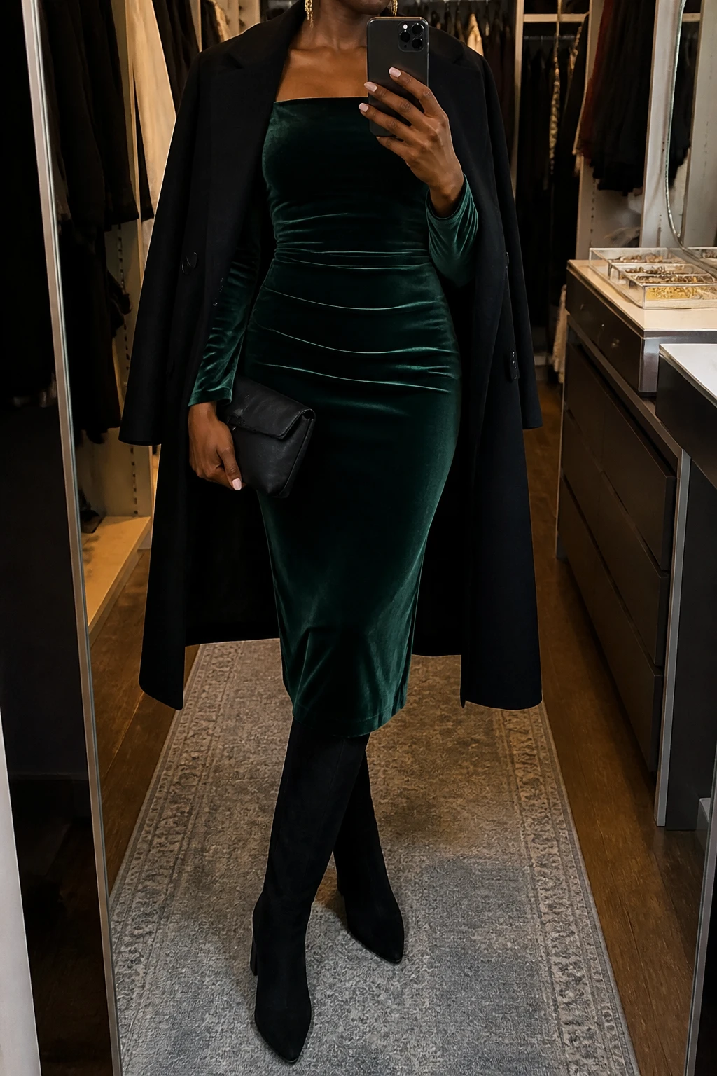 Image Prompt: Mirror selfie of a black woman wearing an emerald green long-sleeve velvet midi dress with a modest square neckline and softly fitted waist, black suede knee-high boots with slim block heels, a long black tailored wool coat worn open, a small black satin evening clutch, and delicate gold drop earrings, holding a phone in front of a mirror, cropped at the NECK so the face does not show, with a relaxed casual selfie pose, in a walk-in closet mirror, warm moody lighting, an open closet with clothes visible and a jewelry tray on the vanity, realistic emerald velvet texture, natural black suede finish, soft evening shadows, latest iPhone photo quality, no additional clothing or accessories.