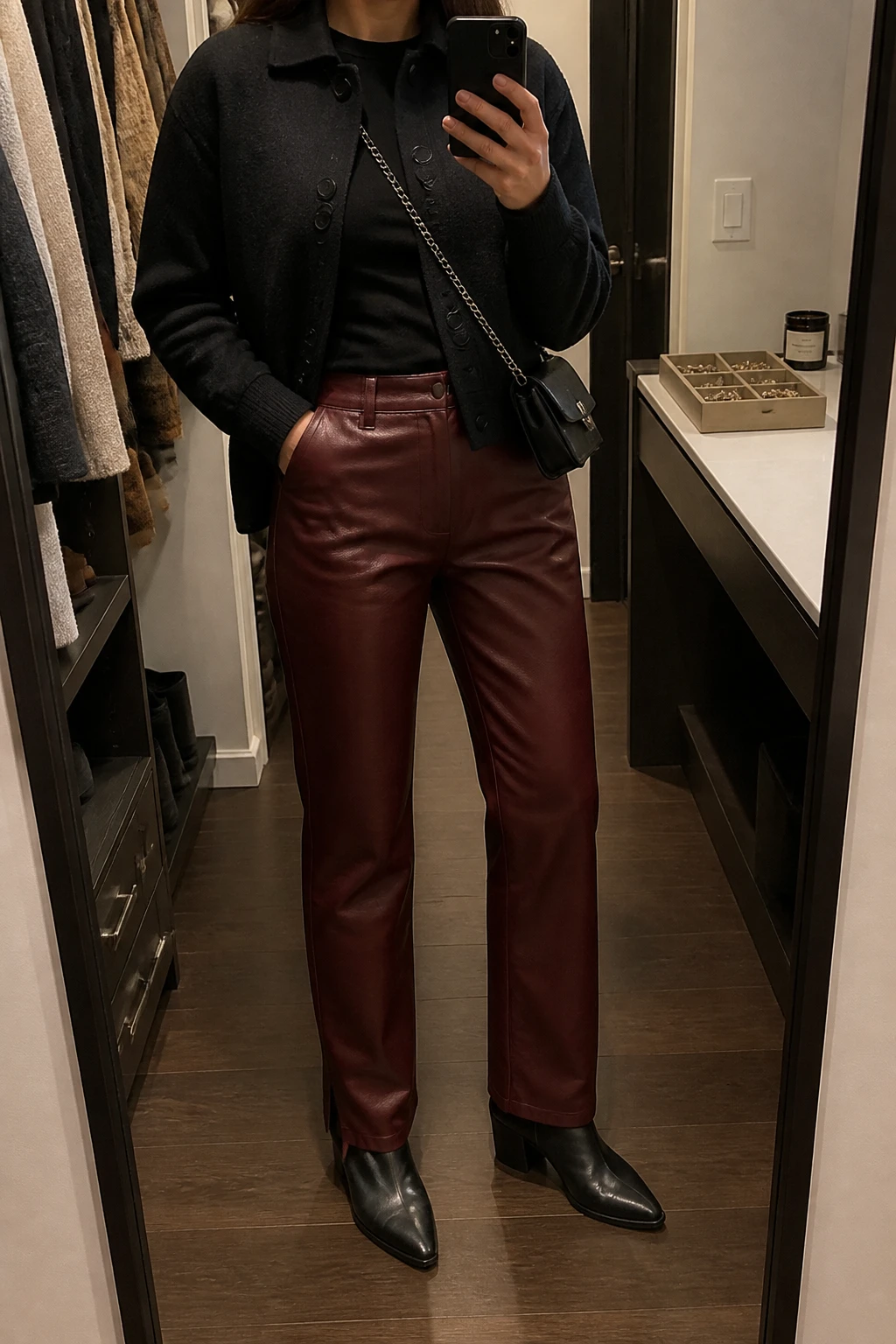 Image Prompt: Mirror selfie of a woman wearing burgundy straight-leg faux-leather trousers, a black fitted long-sleeve stretch jersey top, a cropped black wool-blend jacket, black leather pointed-toe ankle boots with low block heels, and a small black chain-strap crossbody bag, holding a phone in front of a mirror, cropped at the NECK so the face does not show, with a relaxed casual selfie pose, in a walk-in closet mirror, warm moody lighting, an open closet with clothes visible and a jewelry tray on the vanity, natural burgundy leather finish, realistic proportions and subtle fabric wrinkles, latest iPhone photo quality, no additional clothing or accessories.