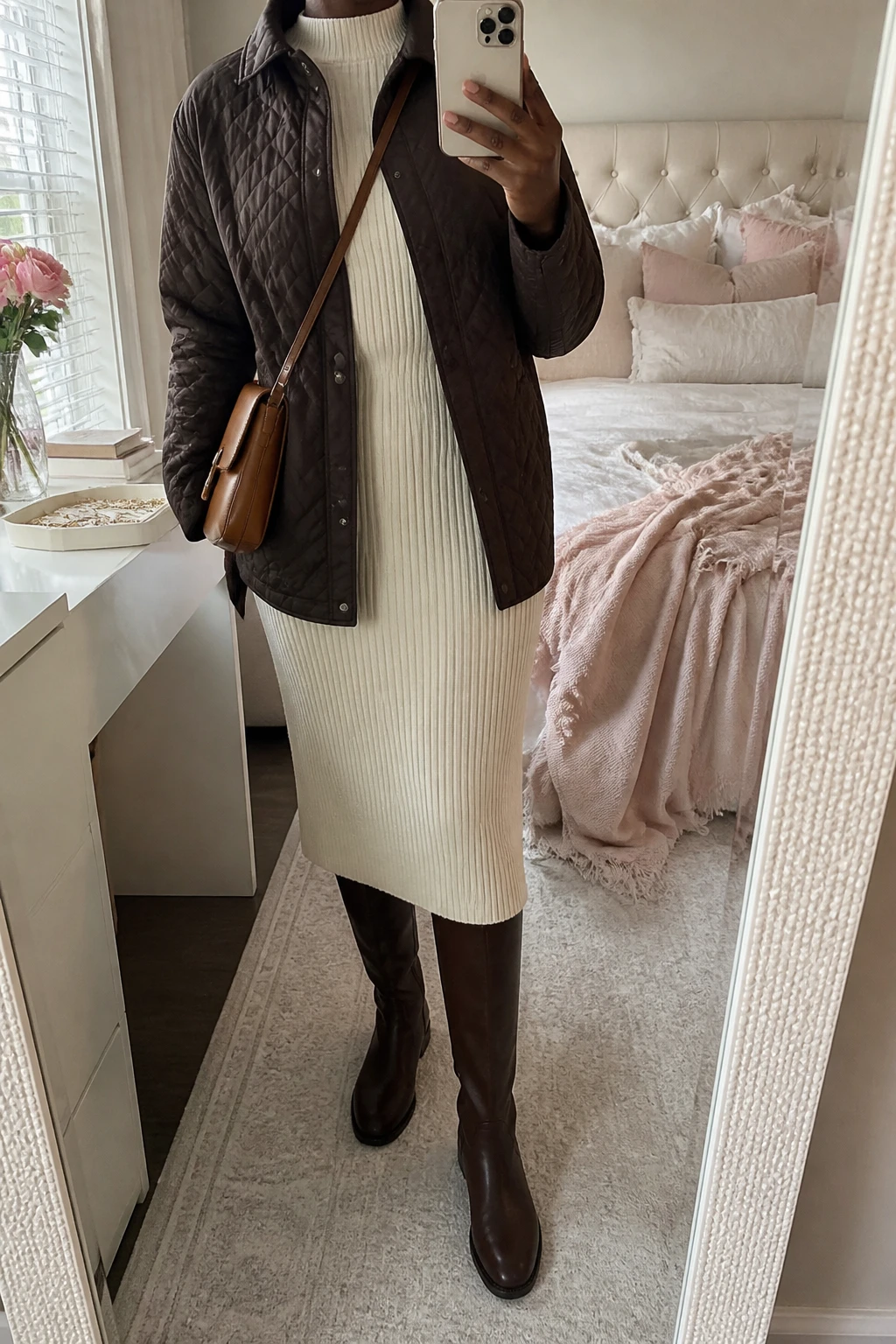 Image Prompt: Mirror selfie of a black woman wearing a chocolate brown insulated quilted hip-length jacket over a cream long-sleeve ribbed knit midi dress with a high neckline and softly fitted waist, dark brown leather knee-high flat boots, a small caramel leather crossbody bag, and delicate gold stud earrings, holding a phone in front of a mirror, cropped at the NECK so the face does not show, with a relaxed casual selfie pose, in a feminine-themed bedroom, soft natural window light, soft pillows on an unmade bed and a jewelry tray on the vanity, realistic chocolate quilted fabric, natural cream ribbed knit folds and soft brown leather texture, latest iPhone photo quality, no additional clothing or accessories.