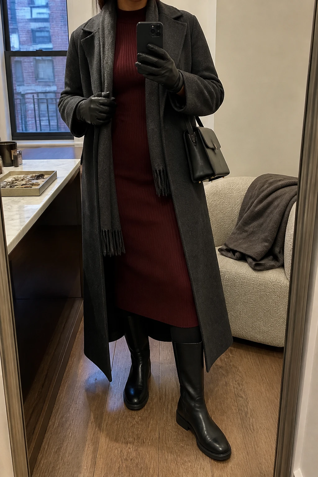 Image Prompt: Mirror selfie of a woman wearing a long charcoal grey heavyweight tailored wool coat worn open over a deep burgundy long-sleeve thick ribbed knit midi dress with a high neckline, black opaque fleece-lined thermal tights underneath, black waterproof leather knee-high flat boots with insulated lining and rubber soles, a charcoal wool scarf, black leather winter gloves, and a small structured black leather shoulder bag, holding a phone in front of a mirror, cropped at the NECK so the face does not show, with a relaxed casual selfie pose, in a stylish city apartment, warm soft indoor lighting, a jewelry tray on the vanity and a folded blanket on a chair, realistic burgundy ribbed dress texture, natural charcoal wool fabric and believable warm winter layers, latest iPhone photo quality, no additional clothing or accessories.