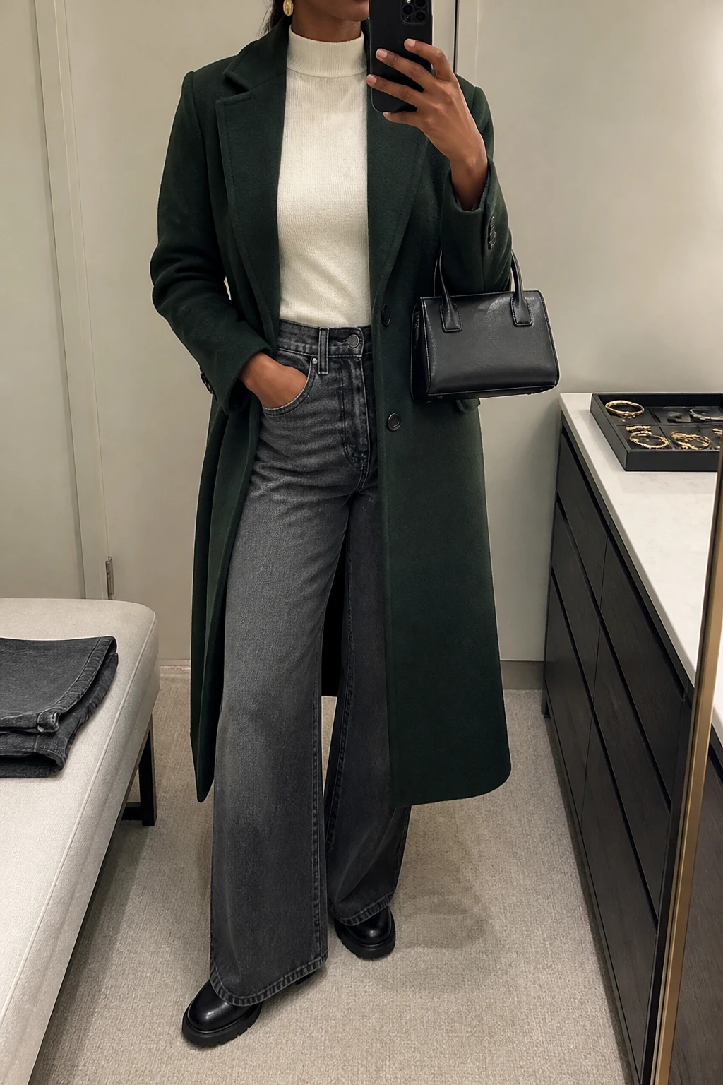Image Prompt: Mirror selfie of a black woman wearing a long forest green single-breasted tailored wool coat worn open over an ivory fitted cashmere mock-neck sweater, charcoal grey high-waisted wide-leg denim jeans with a faded dark-grey wash, black leather chunky flat ankle boots, a small structured black leather handbag, and delicate gold oval earrings, holding a phone in front of a mirror, cropped at the NECK so the face does not show, with a relaxed casual selfie pose, in a minimal dressing room, soft indoor lighting, a jewelry tray on the vanity and folded denim jeans on a bench, realistic forest green wool texture, natural ivory cashmere folds, slightly faded charcoal denim grain, latest iPhone photo quality, no additional clothing or accessories.