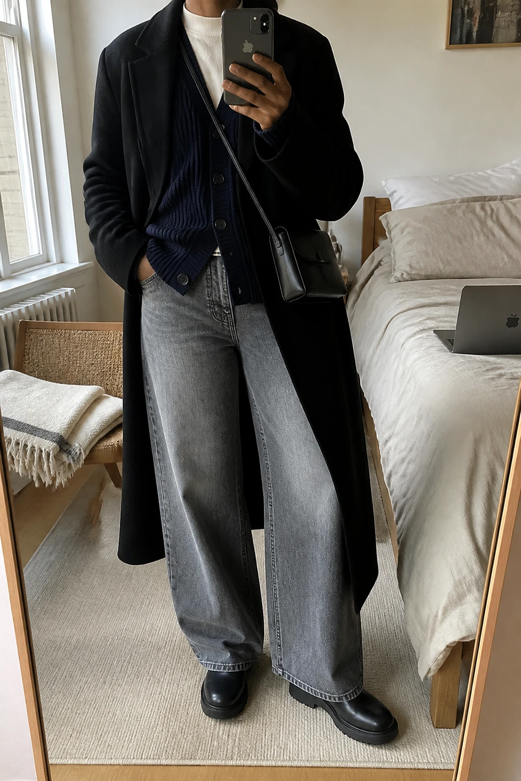Image Prompt: Mirror selfie of a black woman wearing a navy chunky ribbed button-front wool cardigan over an ivory fitted cotton mock-neck top, washed grey high-waisted wide-leg denim jeans, black leather chunky flat Chelsea boots, a long black tailored wool coat worn open, and a small black leather crossbody bag, holding a phone in front of a mirror, cropped at the NECK so the face does not show, with a relaxed casual selfie pose, in a cozy bedroom mirror setup, natural window light, a laptop on the bed and a folded blanket on a chair, realistic navy ribbed knit fabric, natural grey denim wash, black wool coat folds, latest iPhone photo quality, no additional clothing or accessories.