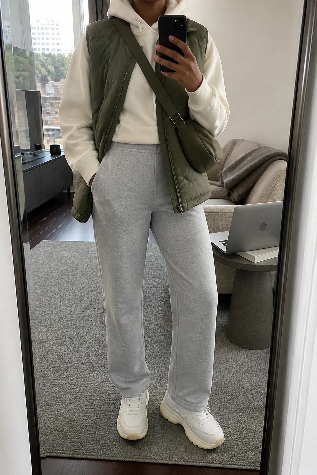 Image Prompt: Mirror selfie of a black woman wearing an olive green insulated quilted puffer vest over a cream oversized fleece-lined hoodie, heather grey high-waisted straight-leg knit joggers, white leather chunky sneakers with beige rubber soles, and a small olive nylon crossbody bag, holding a phone in front of a mirror, cropped at the NECK so the face does not show, with a relaxed casual selfie pose, in a stylish city apartment, soft natural window light, a laptop on a side table and a folded blanket on a chair, realistic olive quilted vest fabric, natural cream hoodie folds and soft grey jogger texture, latest iPhone photo quality, no additional clothing or accessories.