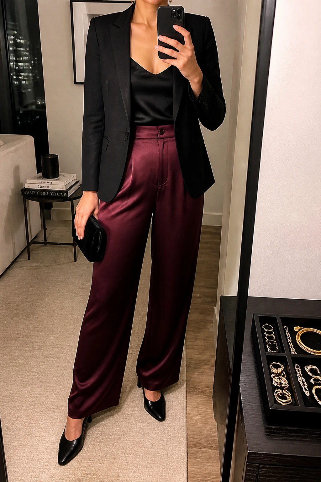 Image Prompt: Mirror selfie of a woman wearing a black fitted single-breasted tailored blazer over a black silk camisole, burgundy high-waisted wide-leg satin trousers, black leather pointed-toe low block heels, a small black velvet clutch, and delicate gold hoop earrings, holding a phone in front of a mirror, cropped at the NECK so the face does not show, with a relaxed casual selfie pose, in a stylish city apartment, warm soft indoor lighting, a jewelry tray on the vanity and a stack of fashion magazines on a side table, realistic satin trouser folds, natural black blazer texture and soft evening shadows, latest iPhone photo quality, no additional clothing or accessories.