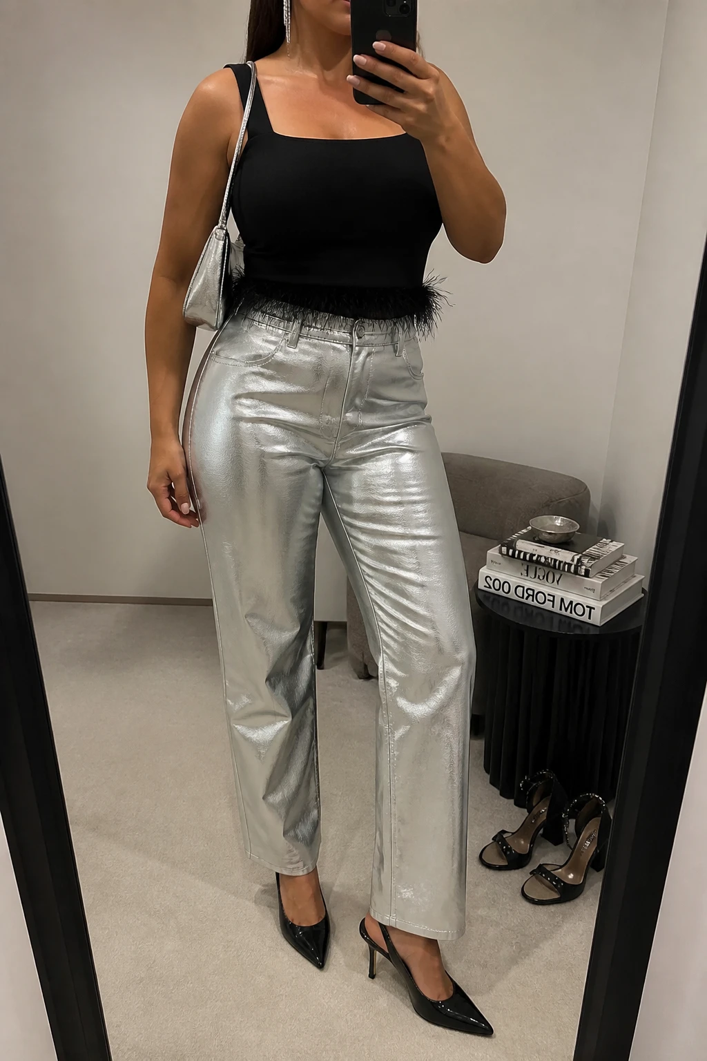 Mirror selfie of a plus size woman wearing silver metallic high-waisted straight-leg trousers, a fitted black sleeveless top with a square neckline and delicate feather trim around the hem, black pointed slingback heels, a small silver leather shoulder bag, and silver linear earrings, clearly showing the exact clothing items, silver trousers, black feather trim, colors, footwear, and accessories listed, holding a phone in front of a mirror, cropped at the neck so her face does not show, relaxed glamorous selfie pose, in a minimal dressing room with soft indoor lighting, a stack of fashion magazines on a side table and a pair of heels on the floor nearby, realistic rooftop birthday styling, latest iPhone photo quality.