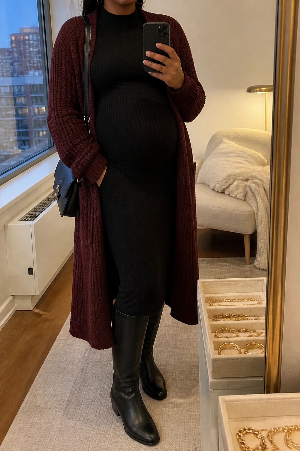 Image Prompt: Mirror selfie of a visibly pregnant black woman wearing a black long-sleeve ribbed knit maternity midi dress with a high neckline and stretchy fabric gently shaping around her baby bump, a long burgundy chunky-knit open-front cardigan, black leather knee-high flat boots, a small structured black leather shoulder bag, and delicate gold hoop earrings, holding a phone in front of a mirror, cropped at the NECK so the face does not show, with a relaxed casual selfie pose and realistic pregnancy proportions, in a stylish city apartment, warm soft indoor lighting, a jewelry tray on the vanity and a folded blanket on a chair, realistic black ribbed knit texture, natural burgundy cardigan stitches, comfortable elegant maternity styling, latest iPhone photo quality, no additional clothing or accessories.